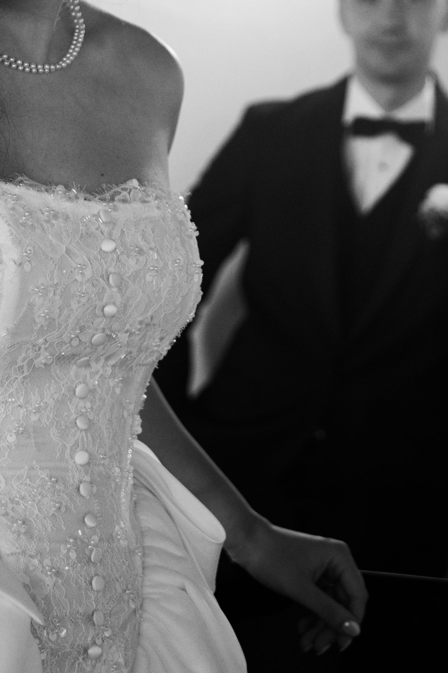 Black and white wedding photo of bride in lace gown with pearl necklace, groom in tuxedo with bow tie in background.