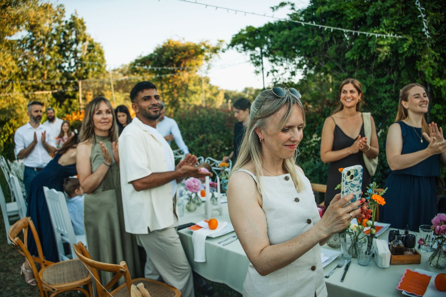 Wedding guests applaud and take photos at an outdoor garden reception with string lights and floral table decor.