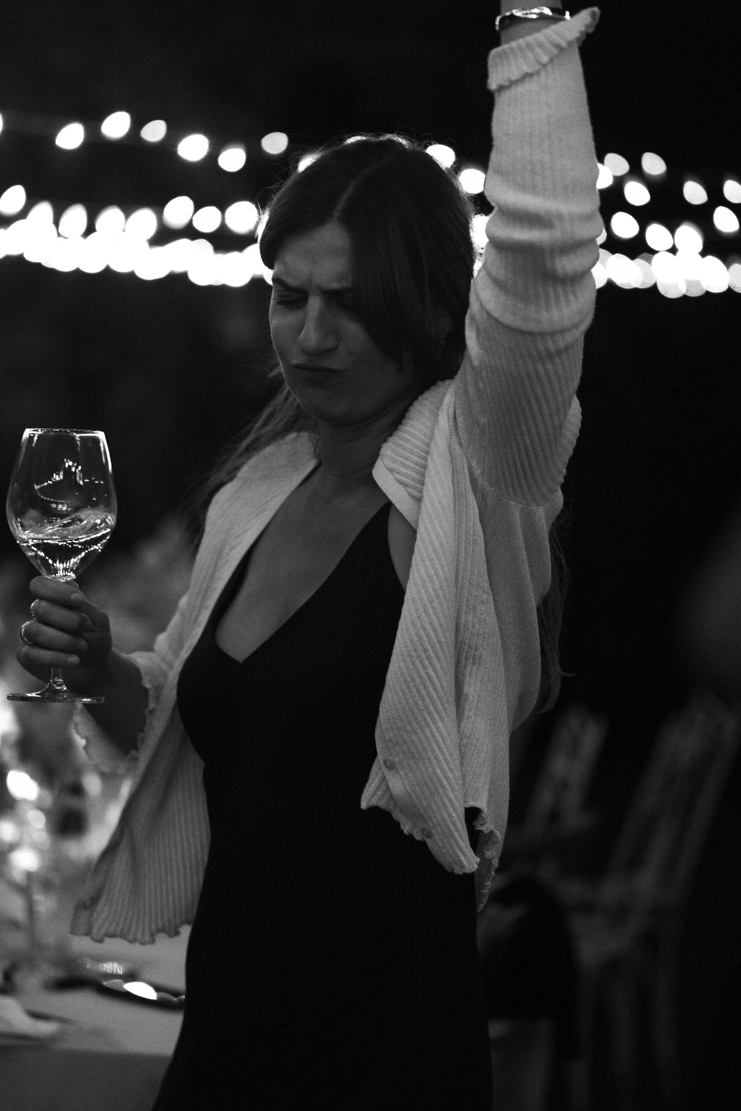 Black and white photo of woman dancing with raised arm holding wine glass at night event.