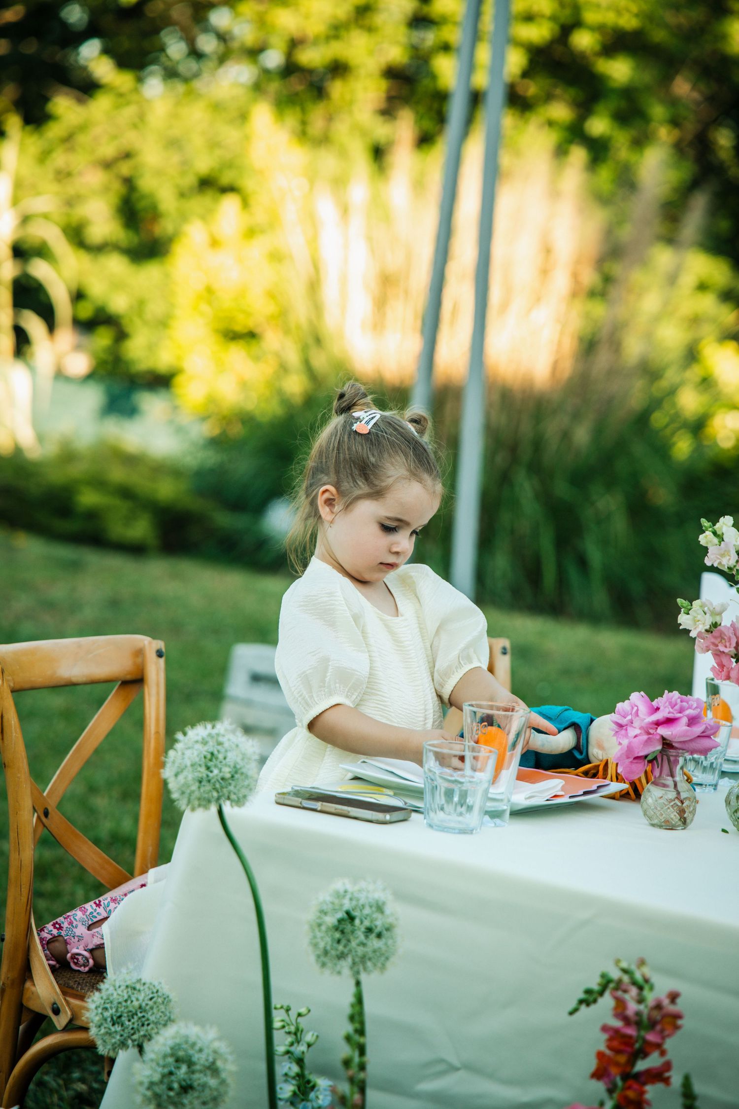 Young girl in white dress sitting at outdoor garden party table decorated with flowers and colorful place settings.