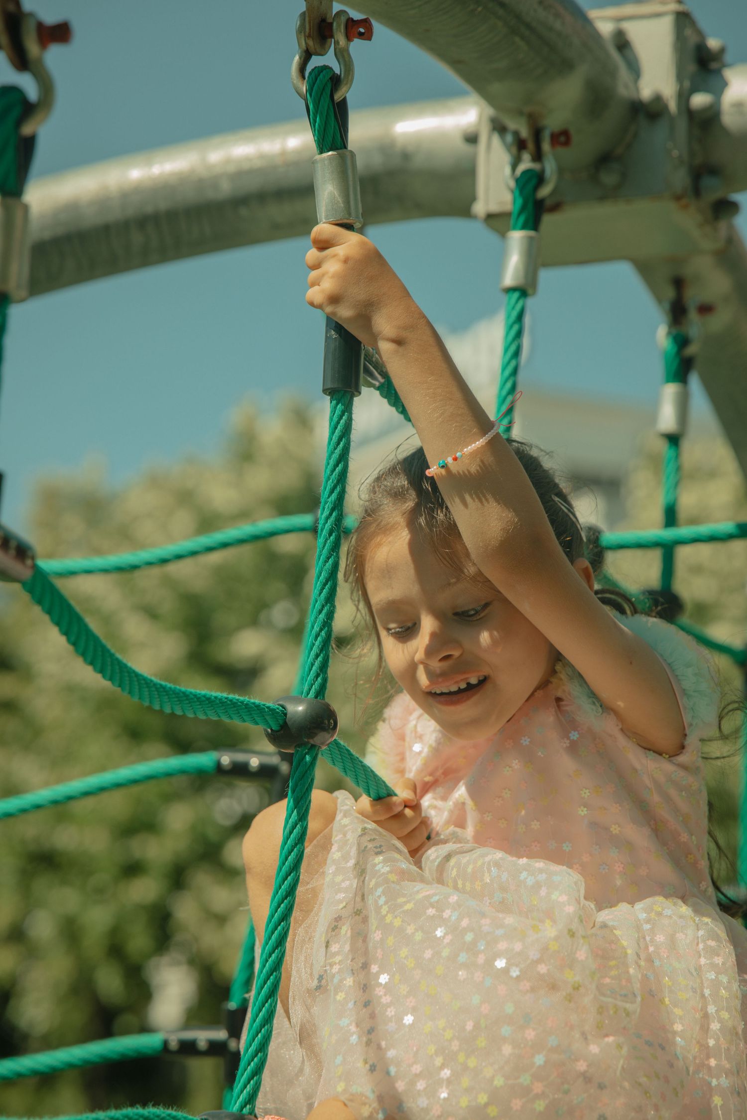 Young girl in sparkly dress climbing green rope playground equipment outdoors on a sunny day.