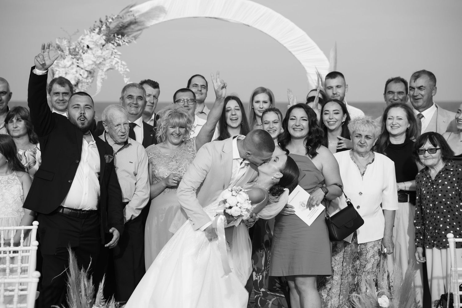 Wedding party celebrates with kiss under floral arch in black and white outdoor ceremony photo.