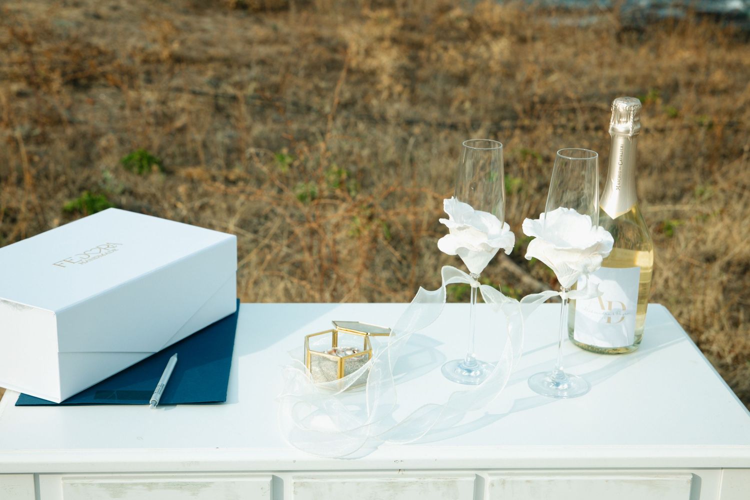 Elegant white table with champagne flutes, white roses, a bottle, and gift boxes for a wedding celebration.