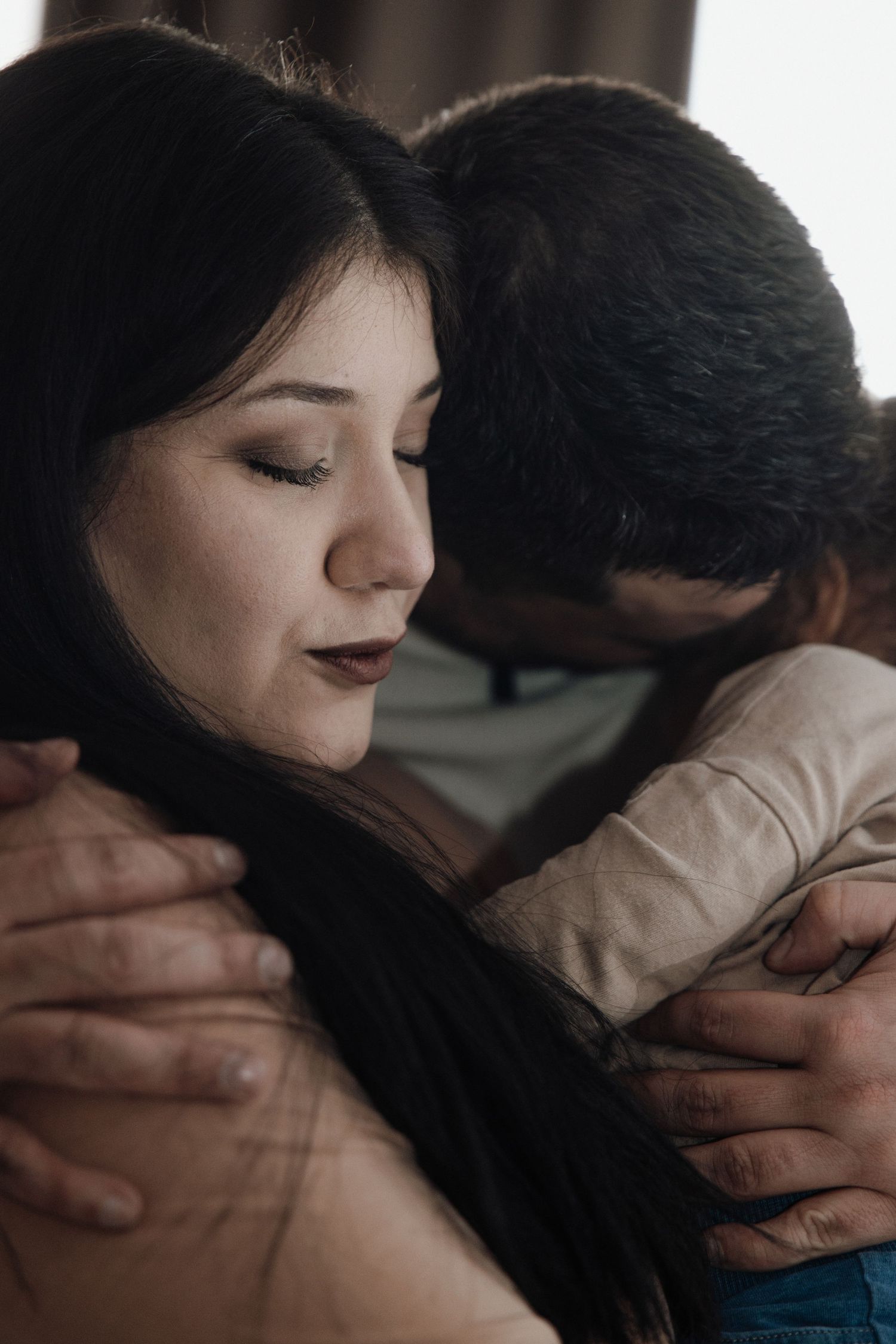 A woman with dark hair closes her eyes while embracing a person, sharing a tender and emotional moment.