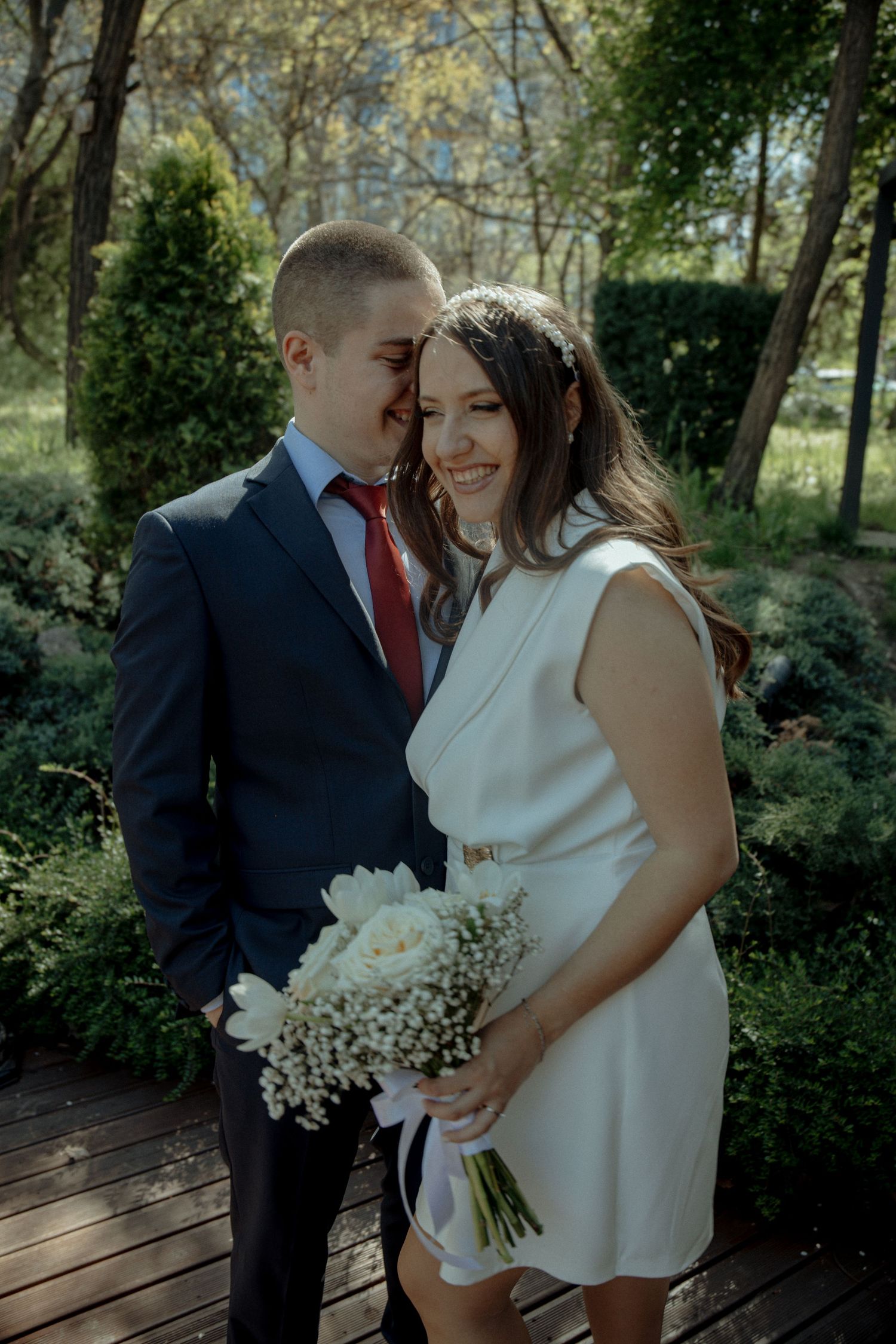 Bride in white dress holding bouquet smiles as groom in navy suit whispers to her in a garden.