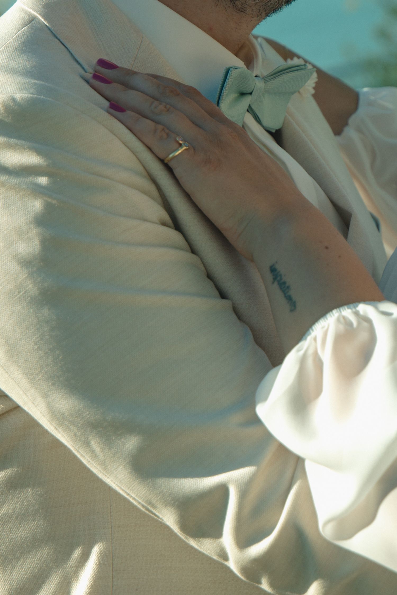 Close-up of bride resting hand on grooms cream suit jacket, wearing gold ring and showing small wrist tattoo.