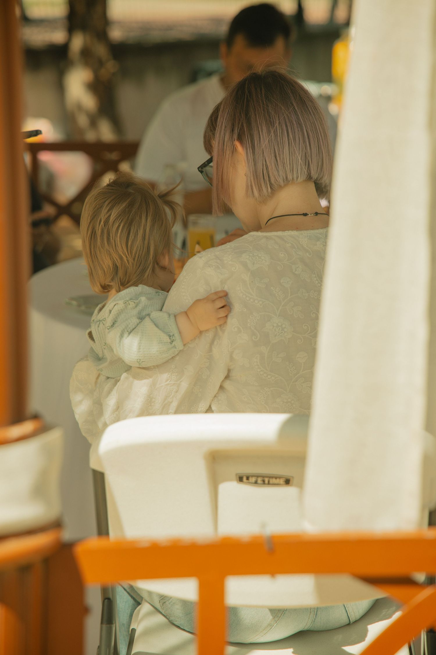 Mother with short blonde hair holding young child at an outdoor seating area.