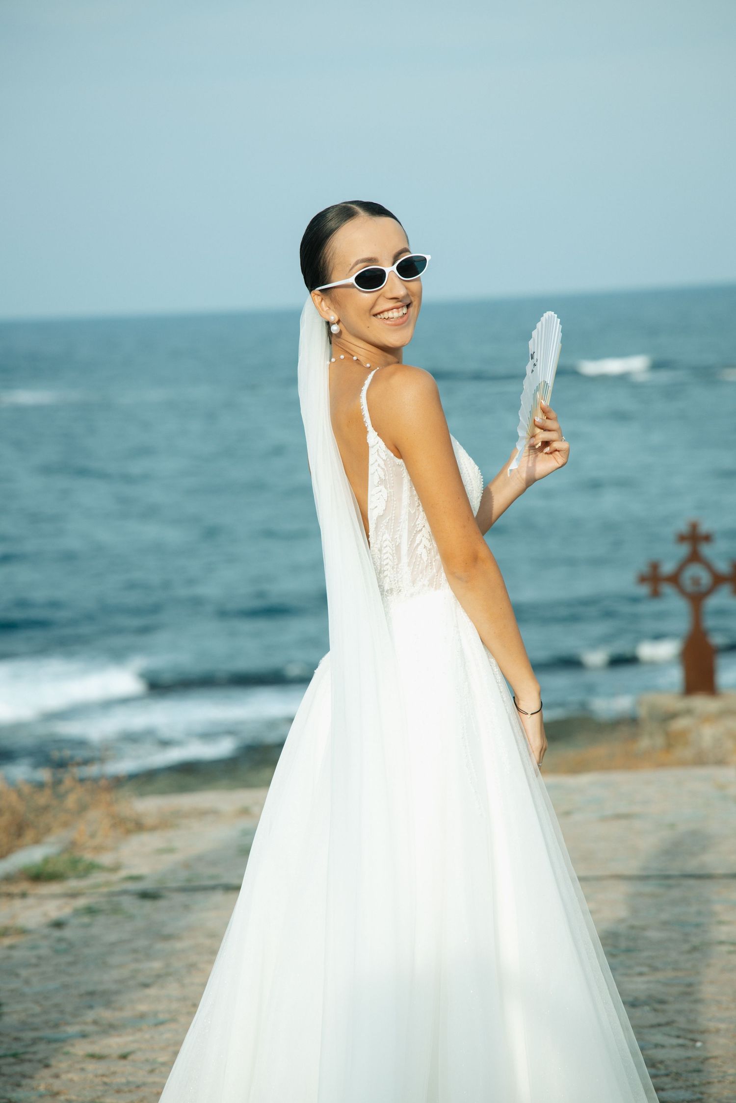 Smiling bride in white wedding gown and sunglasses holding a fan by the ocean.