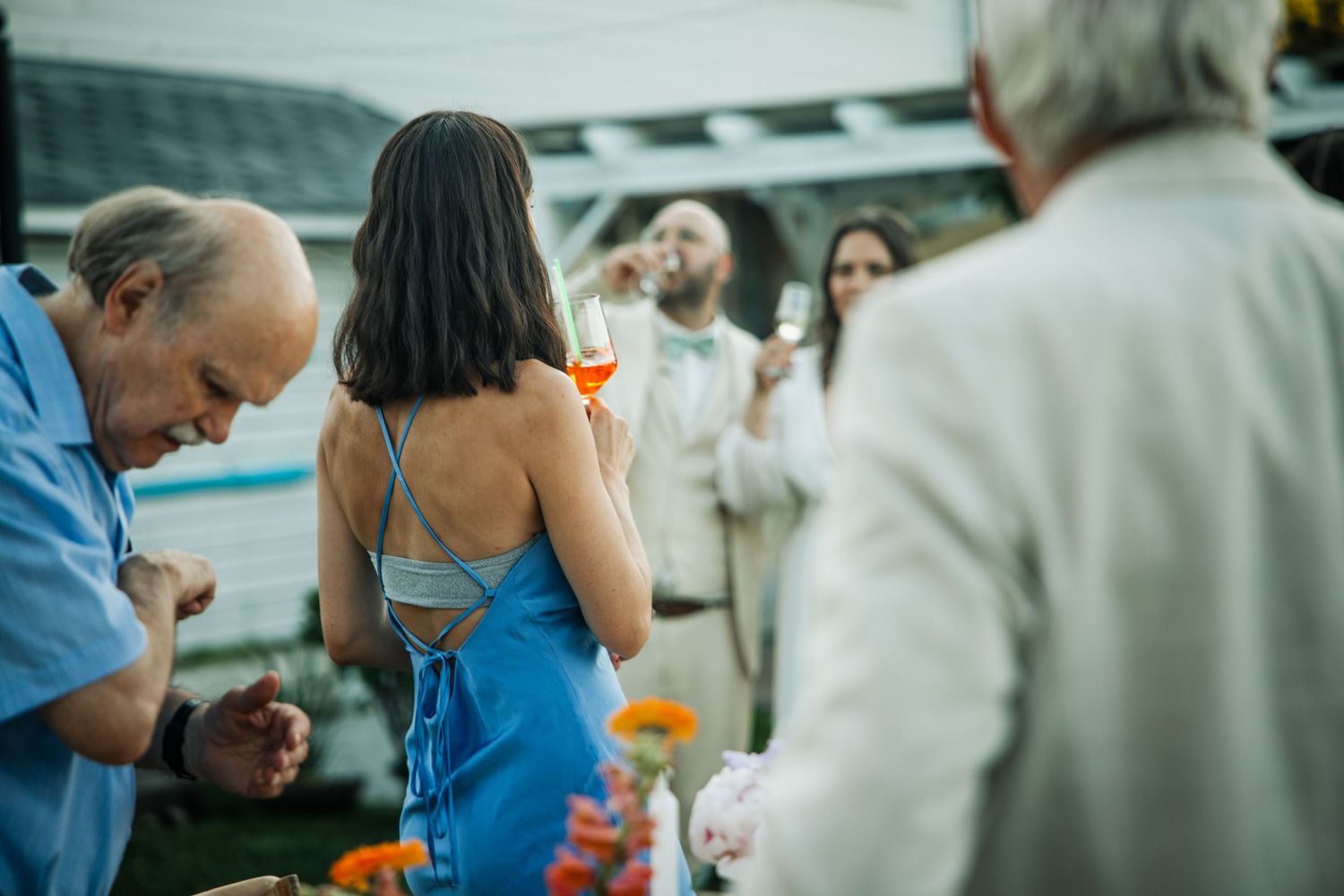 Guests mingle at an outdoor wedding cocktail hour, woman in blue backless dress holds wine glass.
