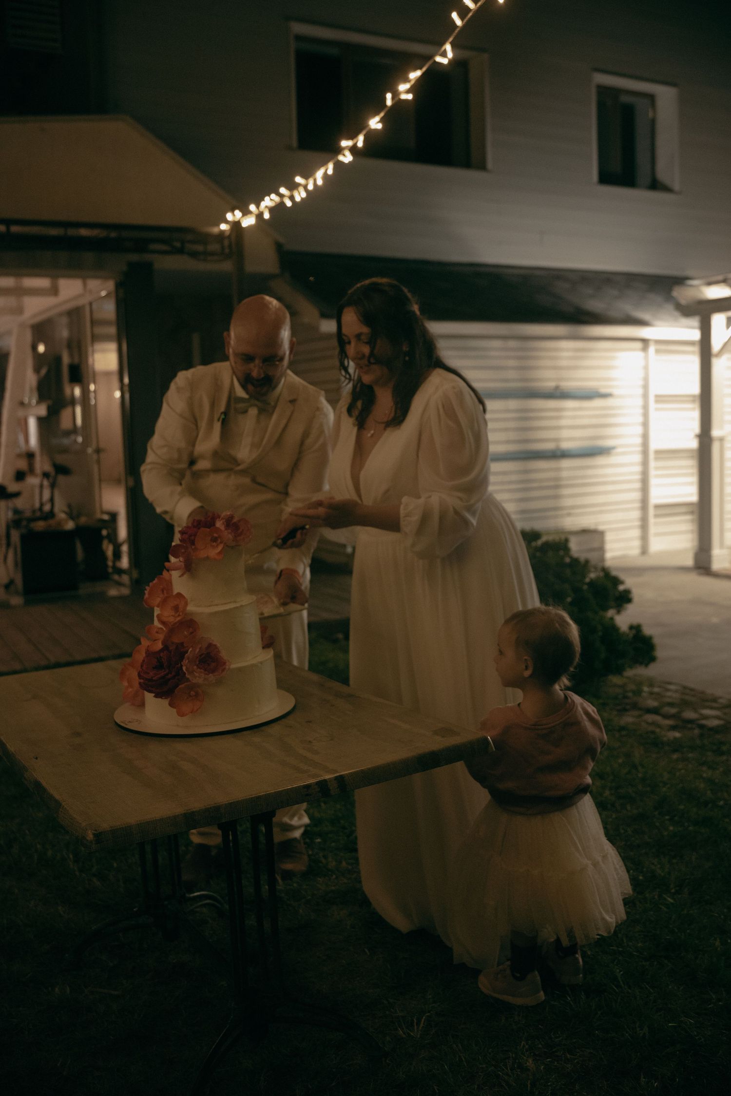 Bride and groom cutting wedding cake outdoors at night with toddler watching under string lights.