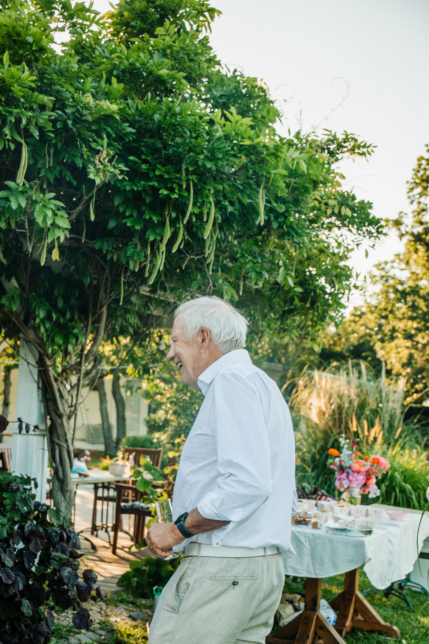 Elderly man in white shirt standing in a lush garden near a decorated wedding reception table.