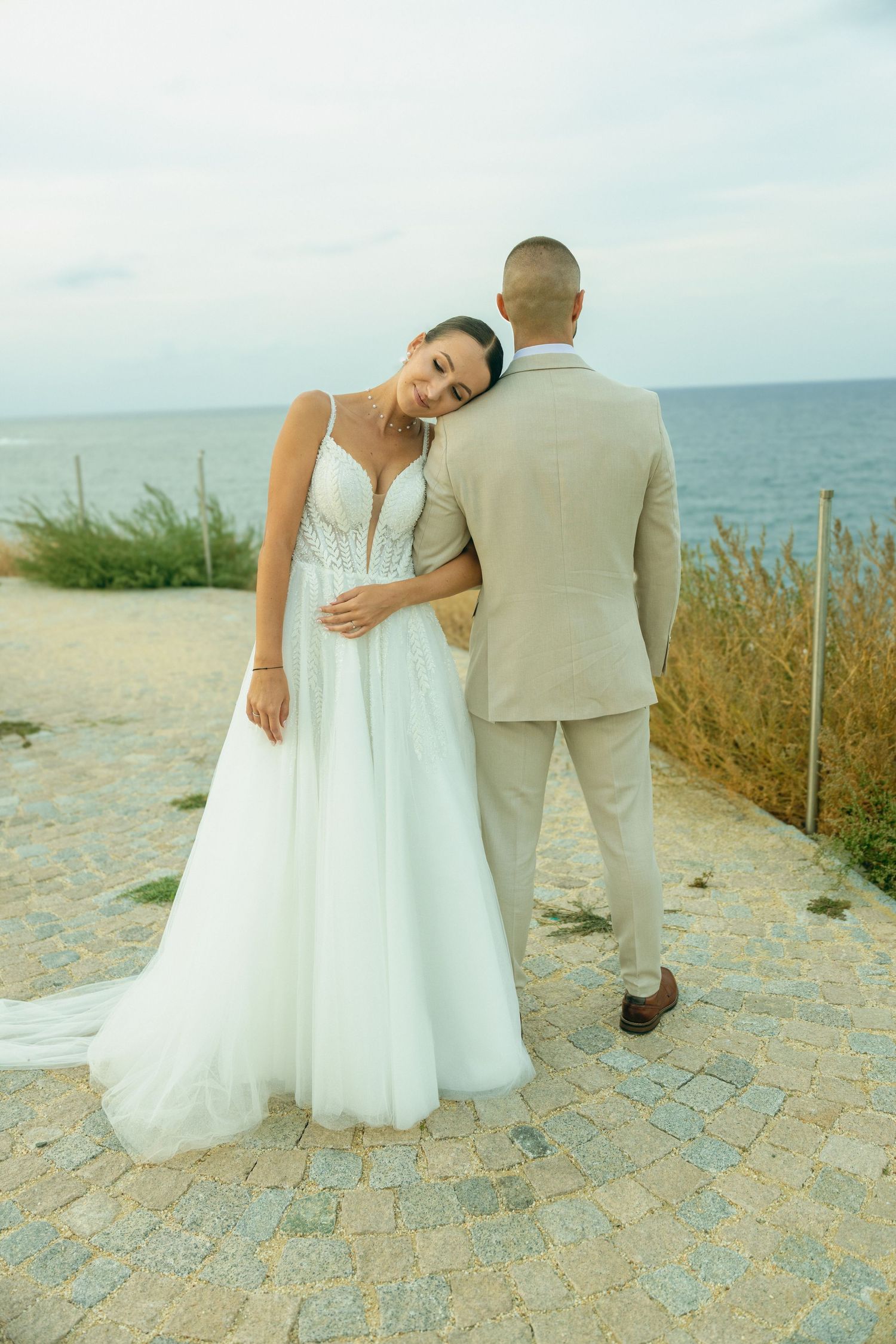 Bride in white gown leans on groom in beige suit by the sea on cobblestone path.