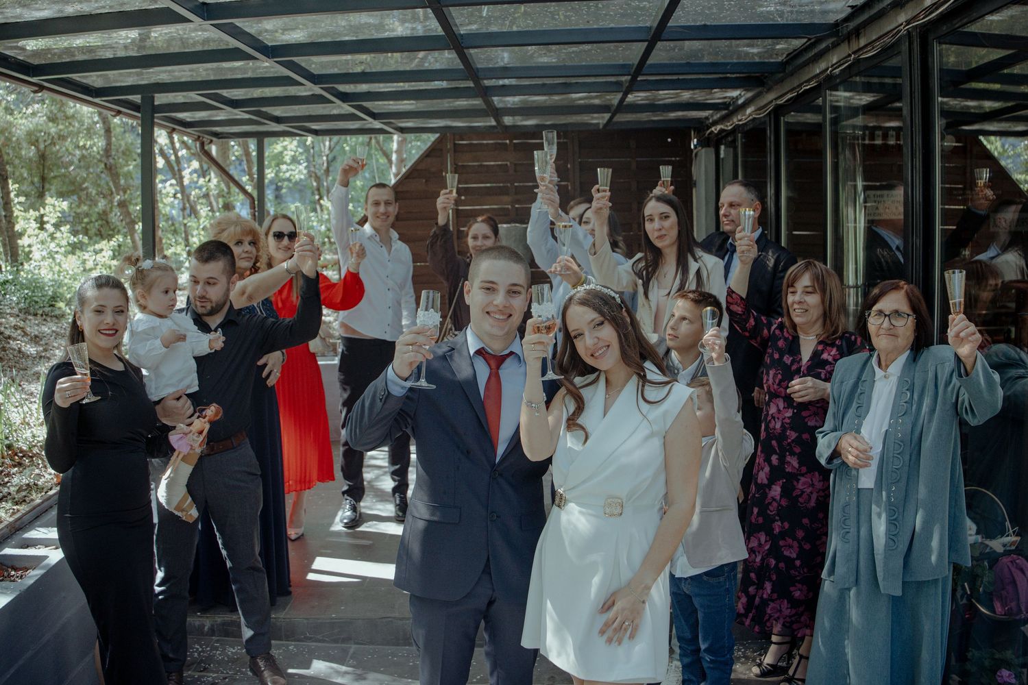 Wedding couple celebrates with guests raising champagne glasses outdoors in a greenhouse venue surrounded by lush greenery.