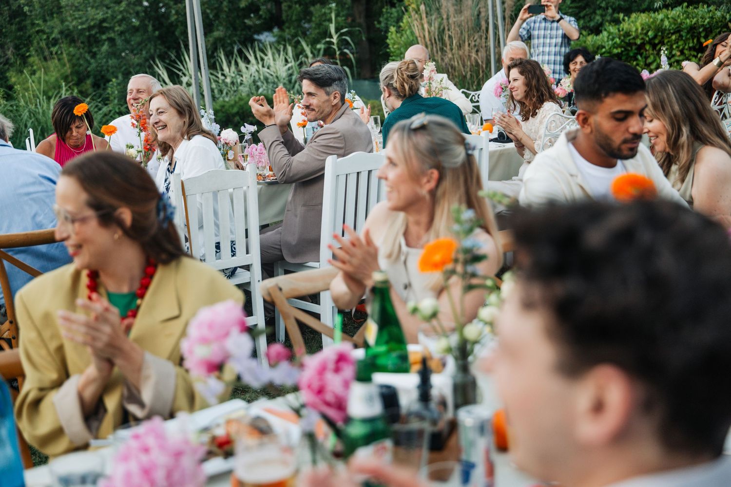Guests socializing and clapping at a lively outdoor wedding reception with floral centerpieces and greenery.