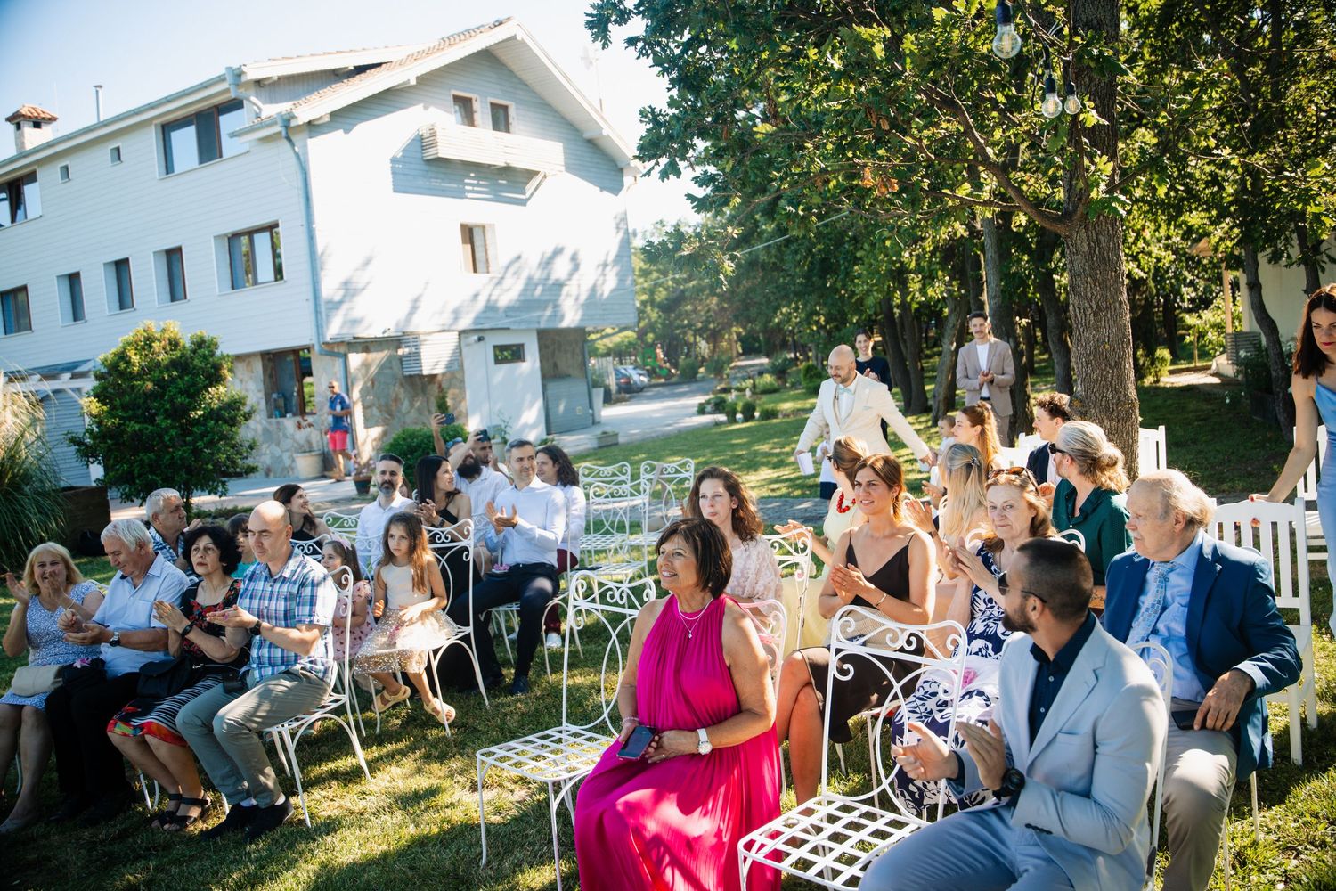 Wedding guests seated on white chairs in a sunny garden with a white building backdrop.