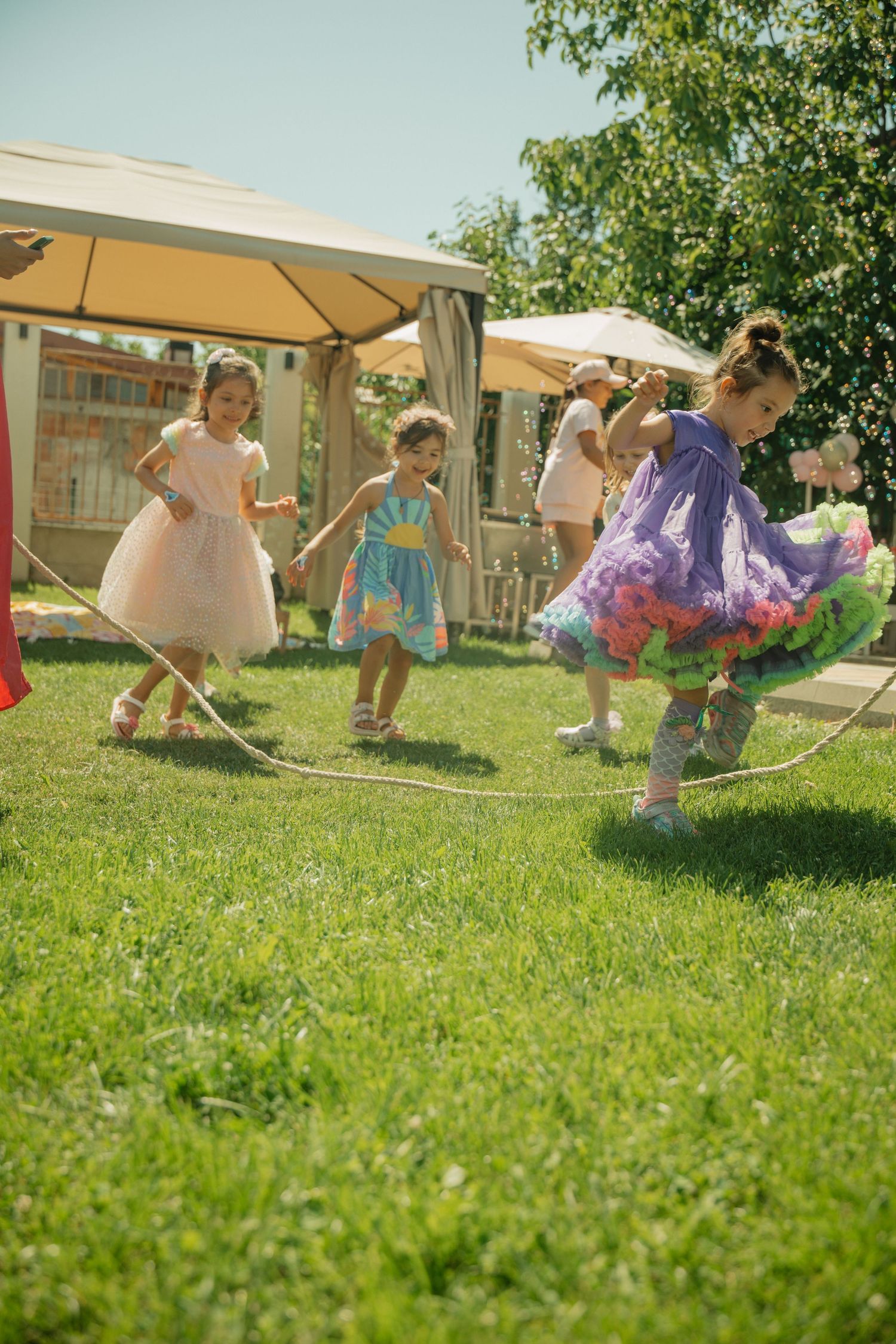 Young girls in colorful dresses jumping rope on green grass at an outdoor party on a sunny day.