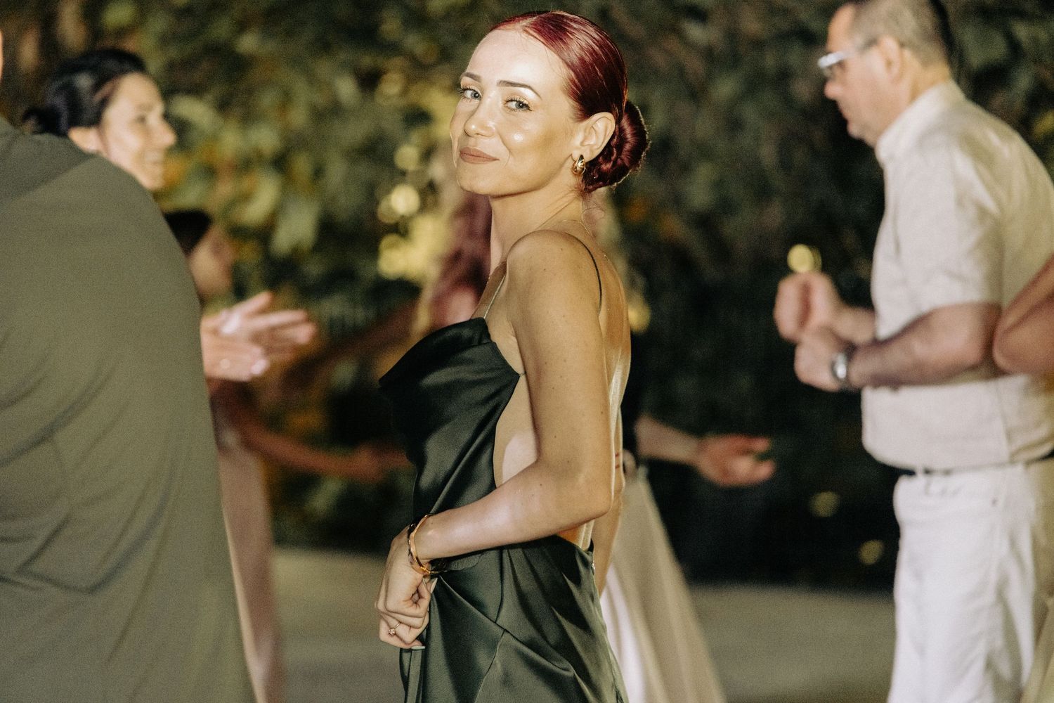 Woman in black satin backless dress smiling at outdoor evening wedding reception party.
