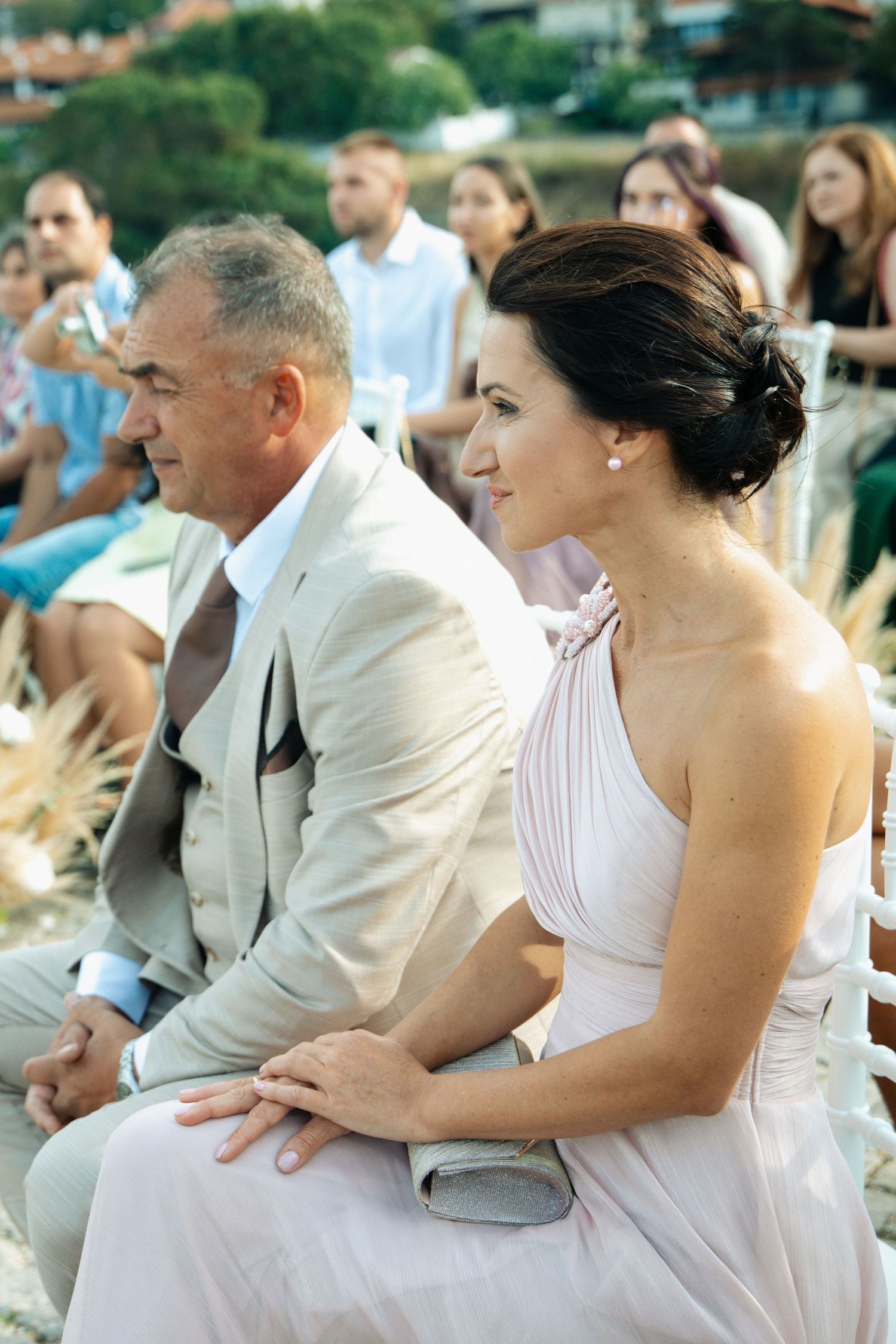 Elegant wedding guests seated outdoors, woman in blush dress and man in beige suit attending a seaside ceremony.
