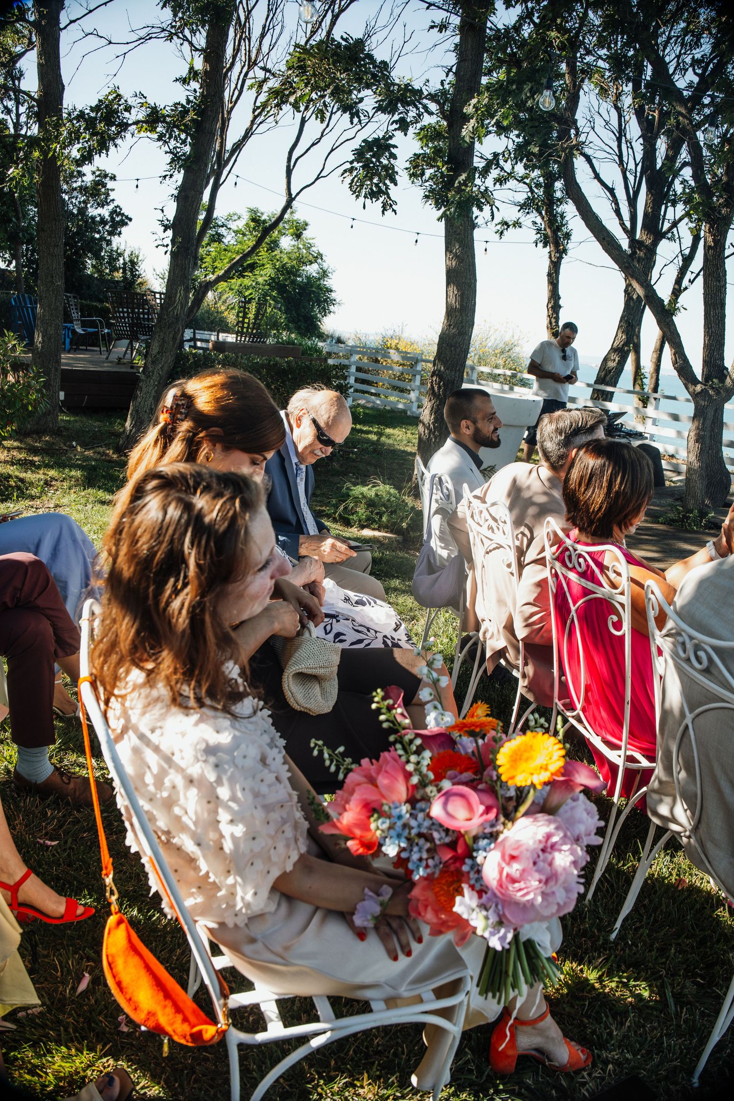 Wedding guests seated on white chairs outdoors holding colorful pink and orange floral bouquet.