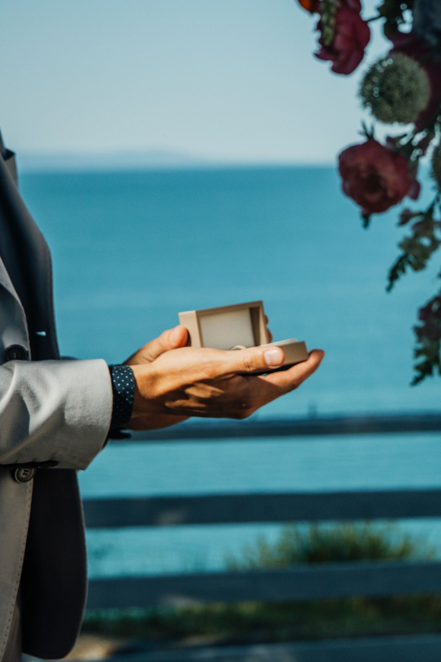 Best man holding open wooden ring box with wedding bands beside a blue ocean backdrop.