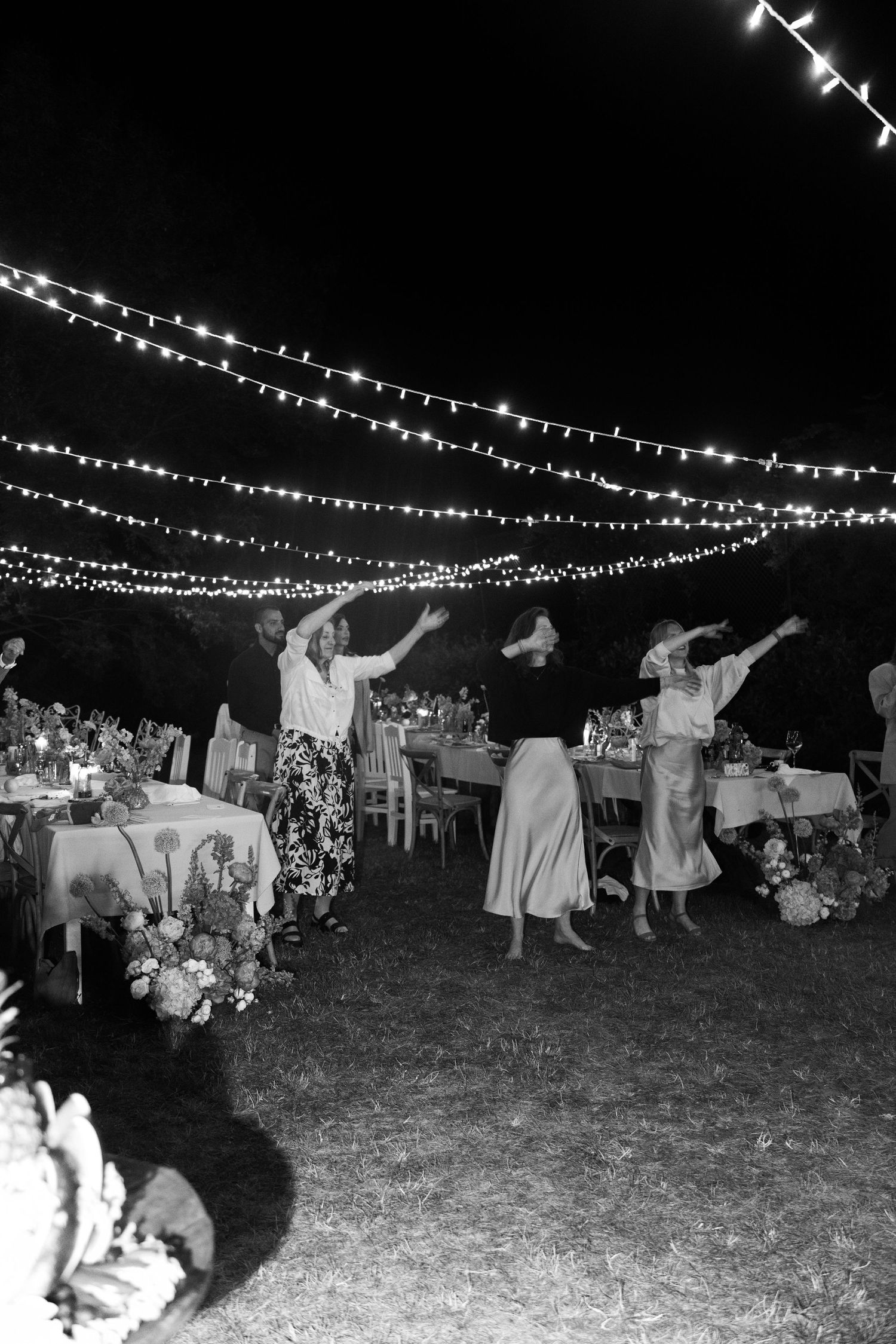 Guests dance joyfully under twinkling string lights at an outdoor nighttime wedding reception in black and white.