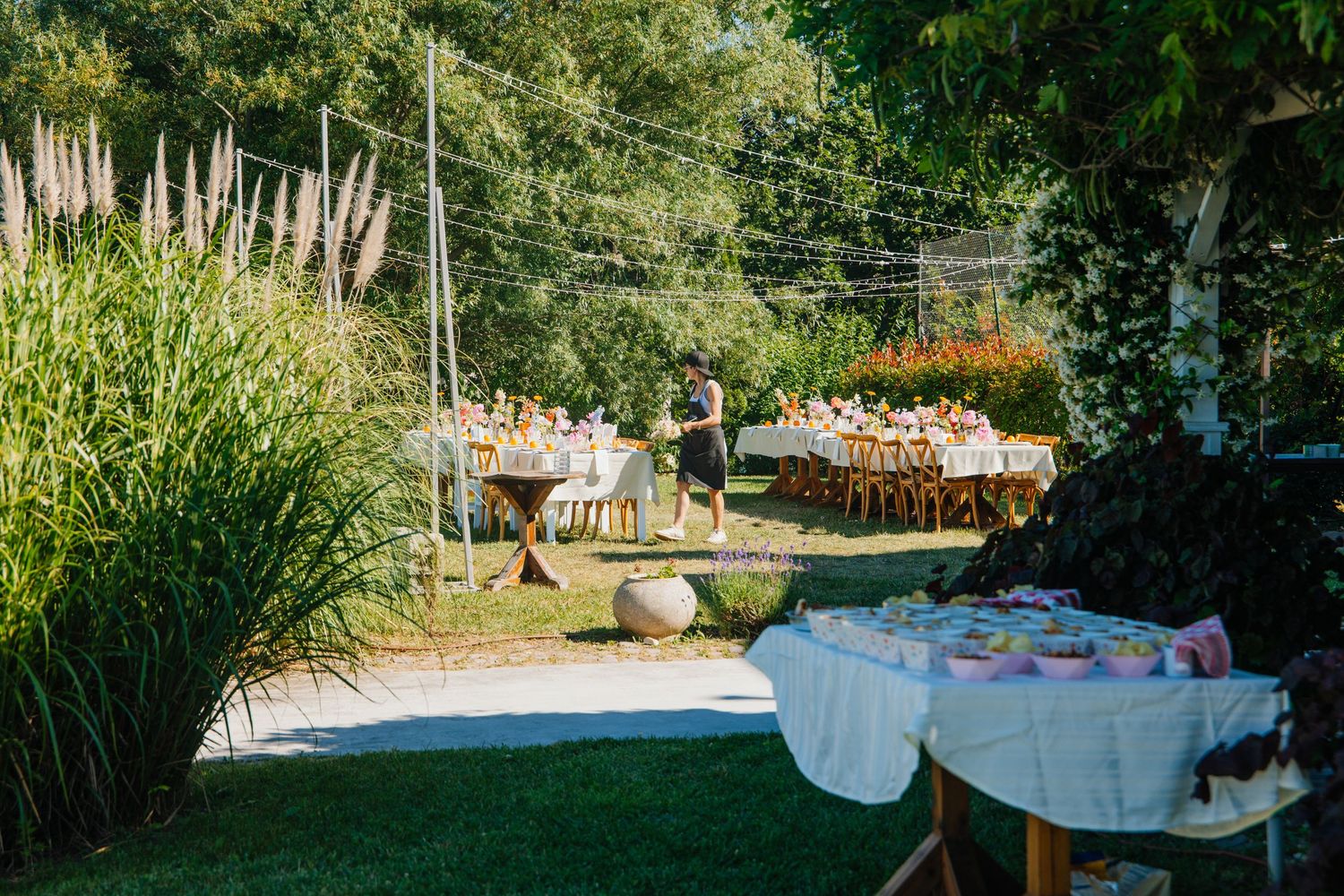 Outdoor garden party setup with long decorated tables, lush greenery, and guests enjoying a sunny afternoon celebration.