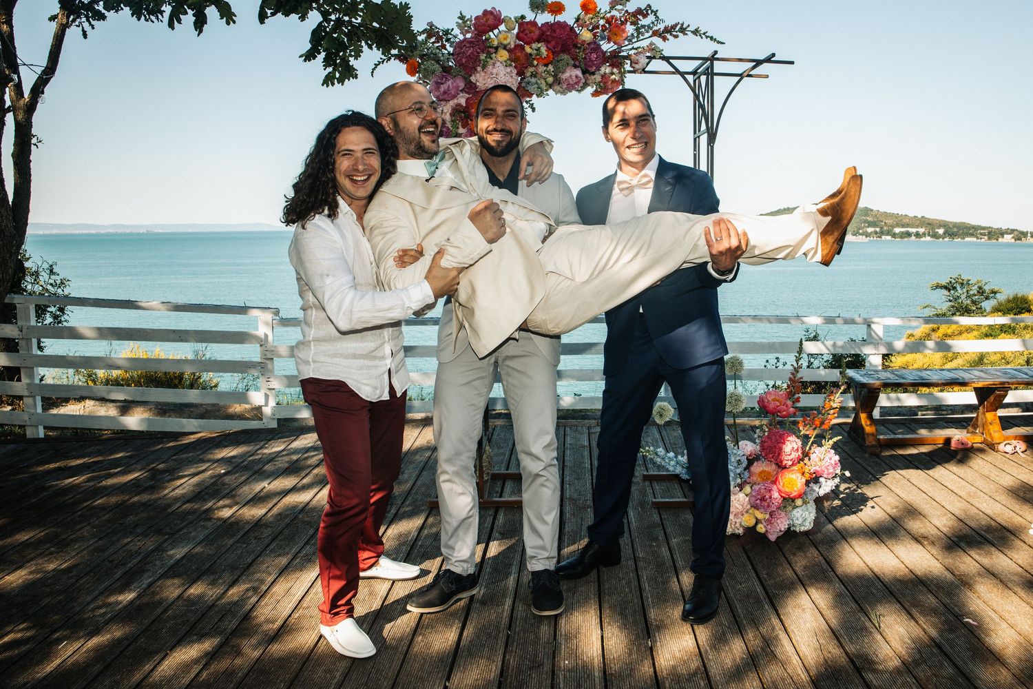 Four groomsmen posing playfully on deck with ocean view at wedding celebration.