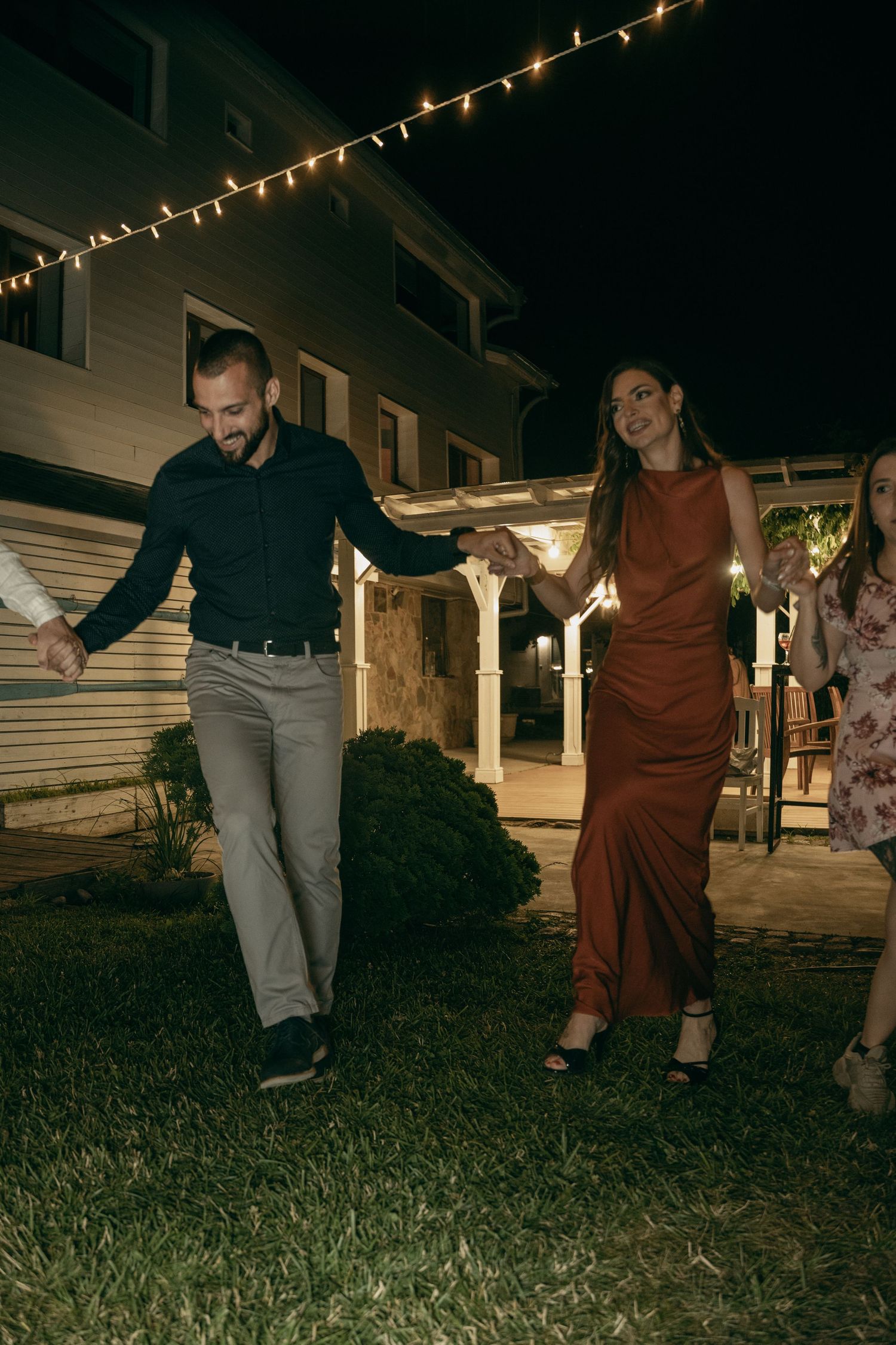 Couple in elegant attire holding hands walking on grass under string lights at night event.