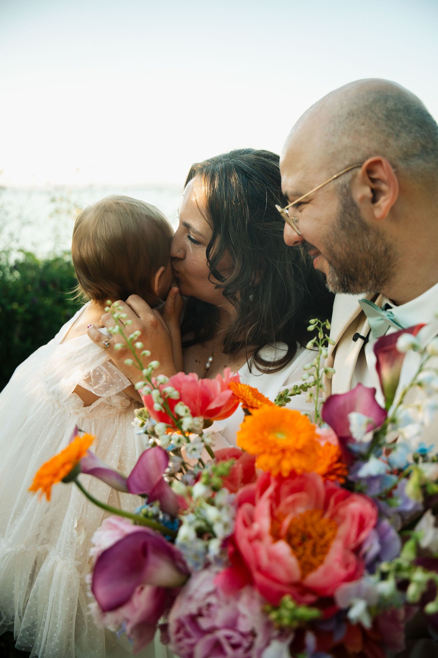 Bride kissing baby while groom smiles, with vibrant colorful wedding bouquet of peonies and flowers in foreground.
