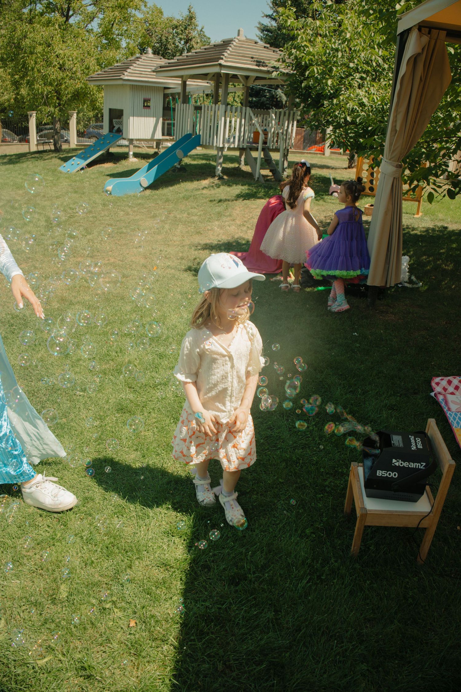 Children in colorful dresses playing with bubbles outdoors at a sunny garden party near a playground.