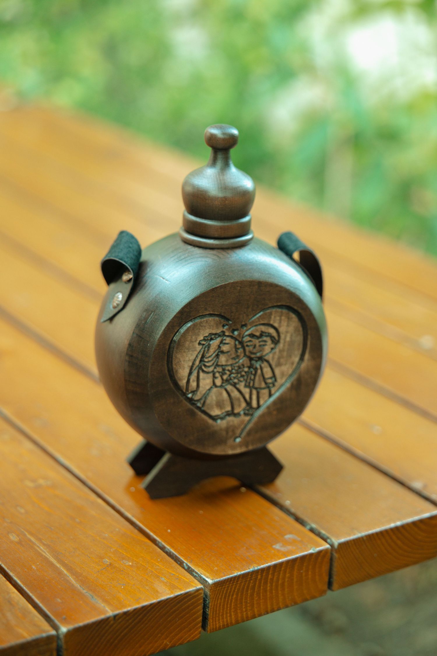 Carved wooden wedding flask with heart-shaped couple engraving displayed on rustic wooden table outdoors.