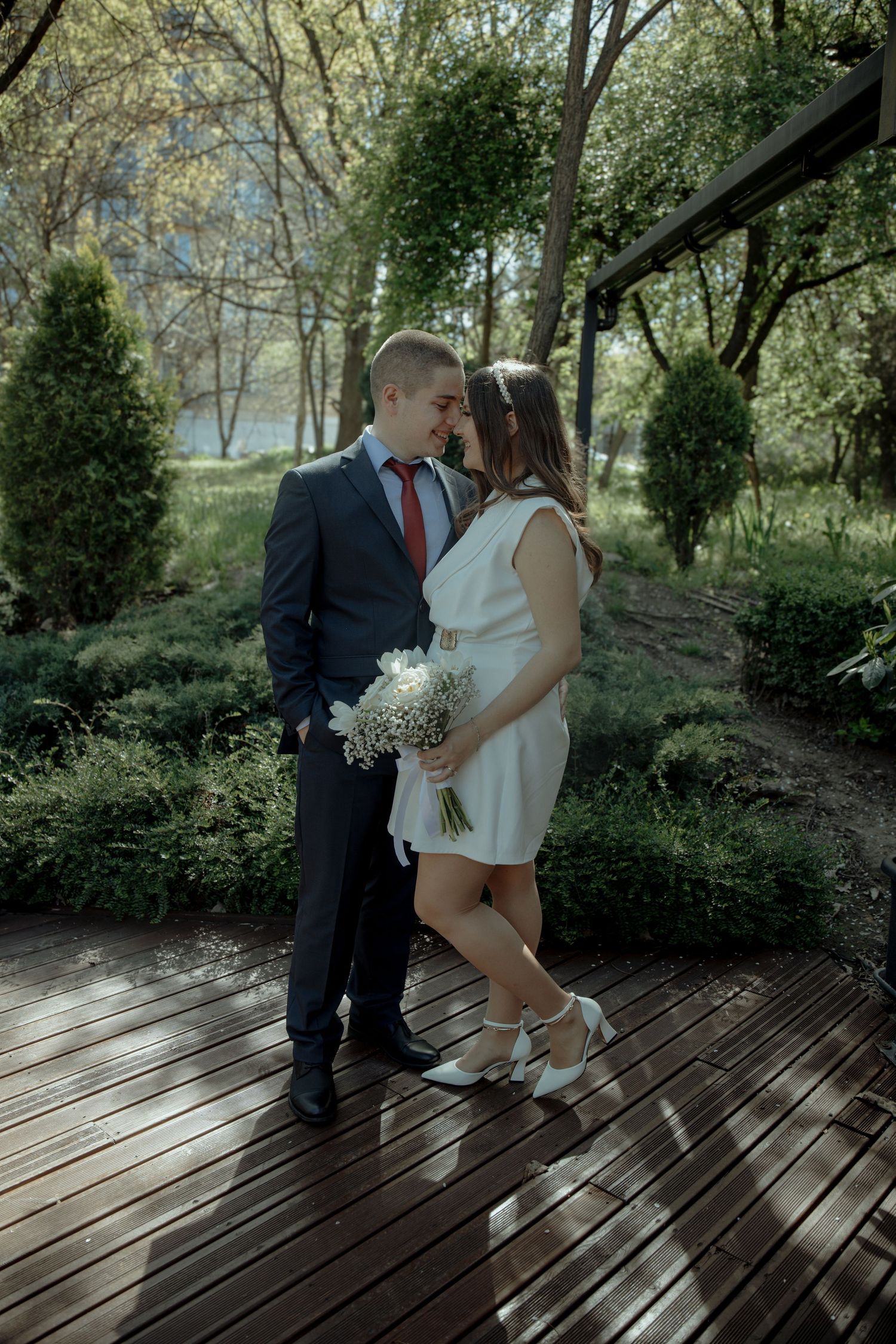 A couple in wedding attire shares a romantic moment on a wooden deck surrounded by lush greenery.