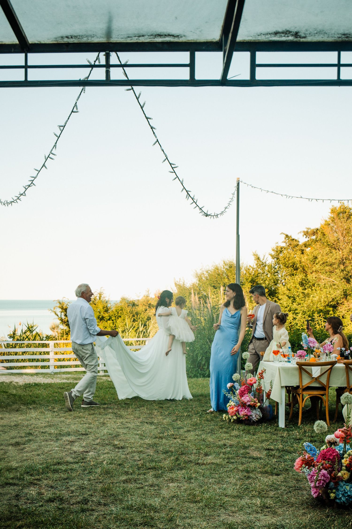 Bride in white gown with wedding party gathered on lawn near seaside at golden hour.