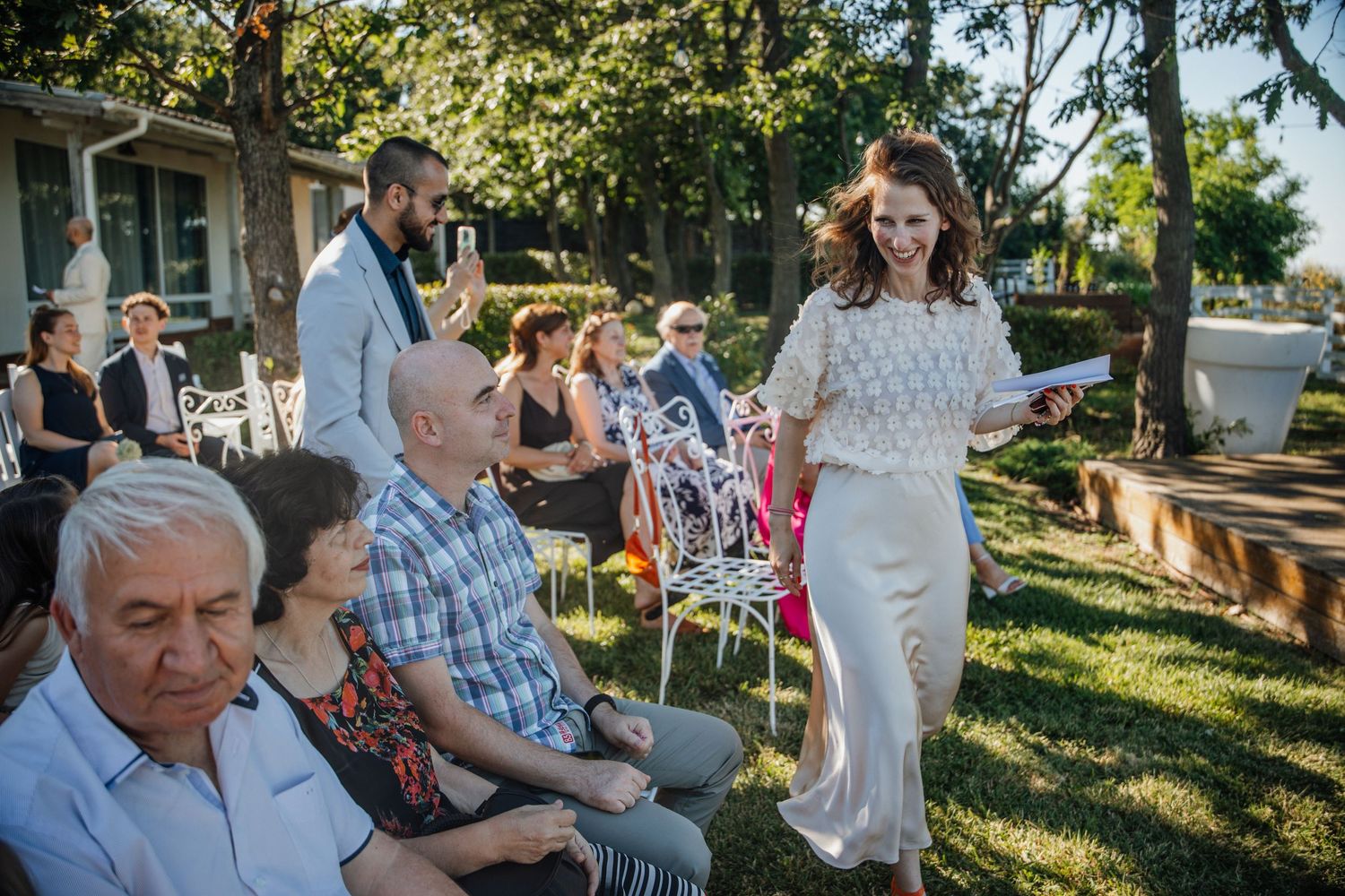Bride in white lace dress walks among seated wedding guests outdoors during a sunny garden ceremony.