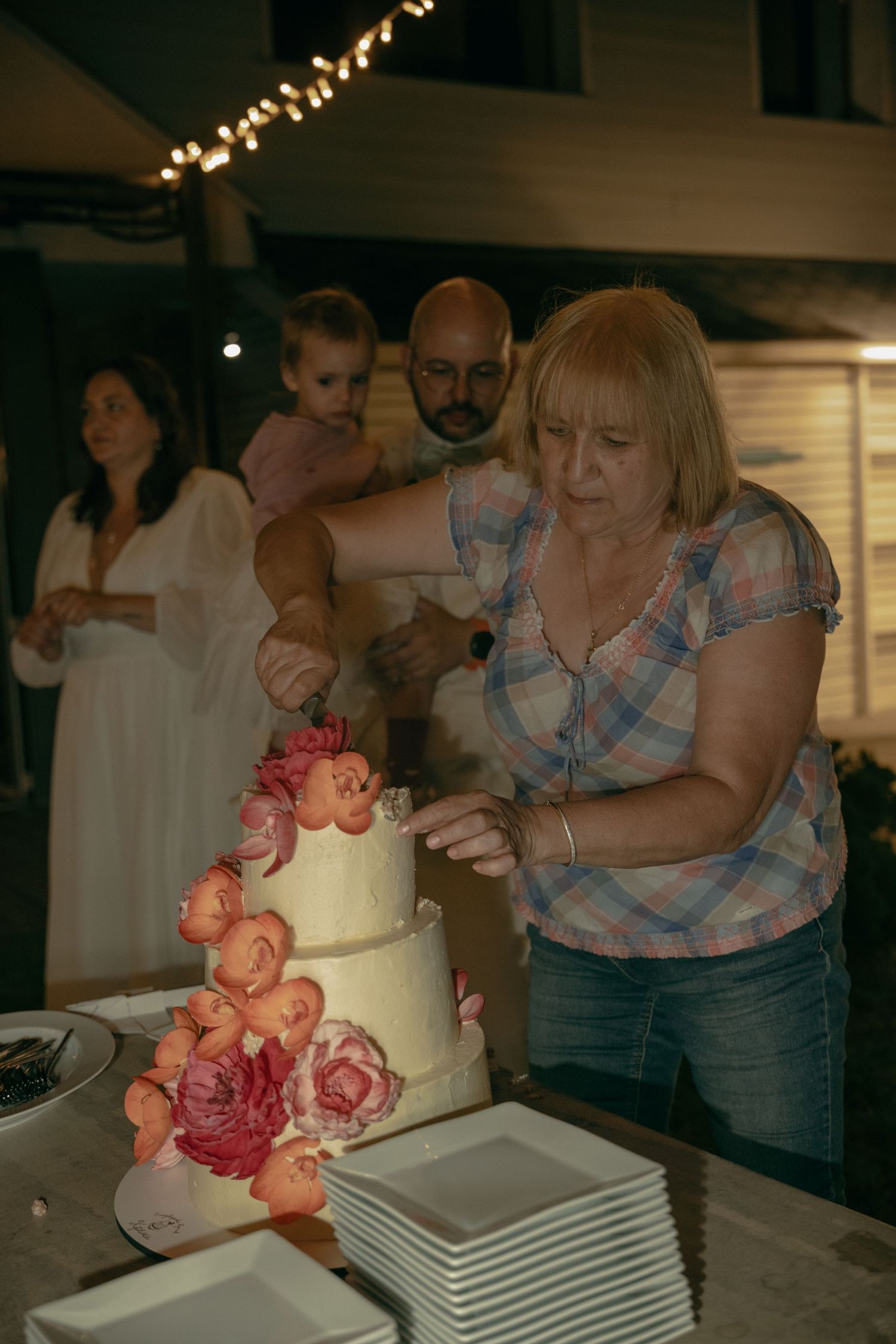 Woman cuts a elegant three-tier floral wedding cake while guests watch at an evening reception.