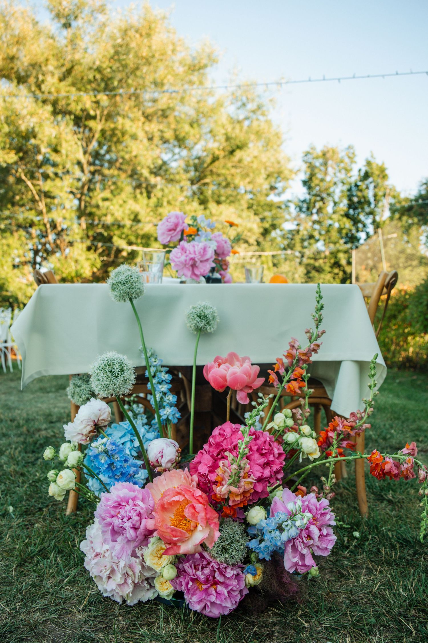 Vibrant garden party floral arrangement with pink peonies, blue hydrangeas, and allium blooms beside an outdoor dining table.