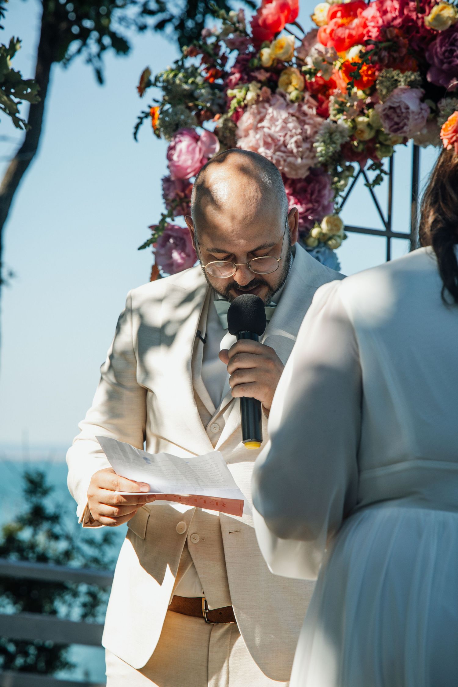 Groom in white suit reads vows at outdoor wedding altar adorned with colorful flowers.