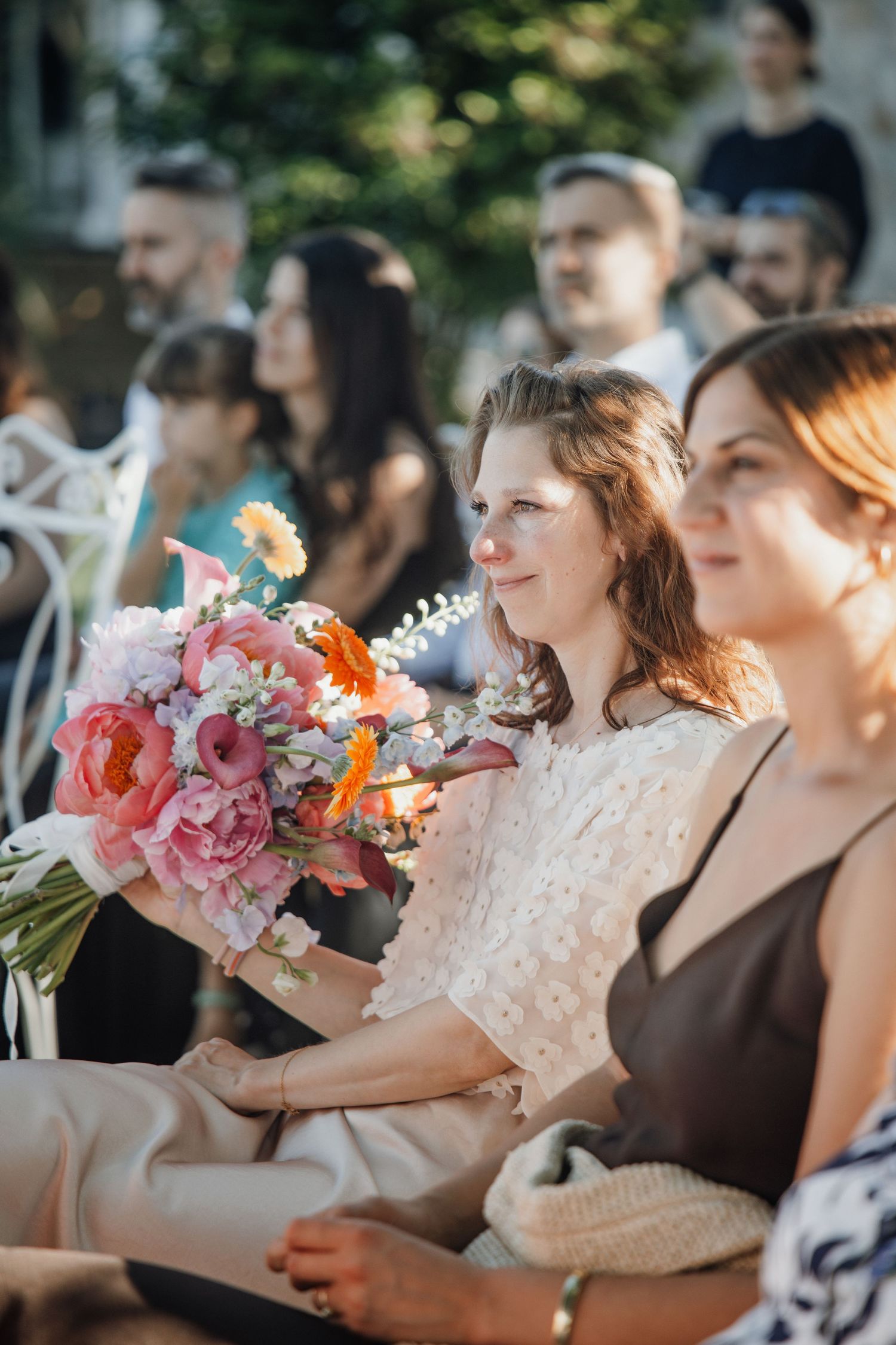 Woman holding colorful pink and orange bridal bouquet seated at outdoor wedding.