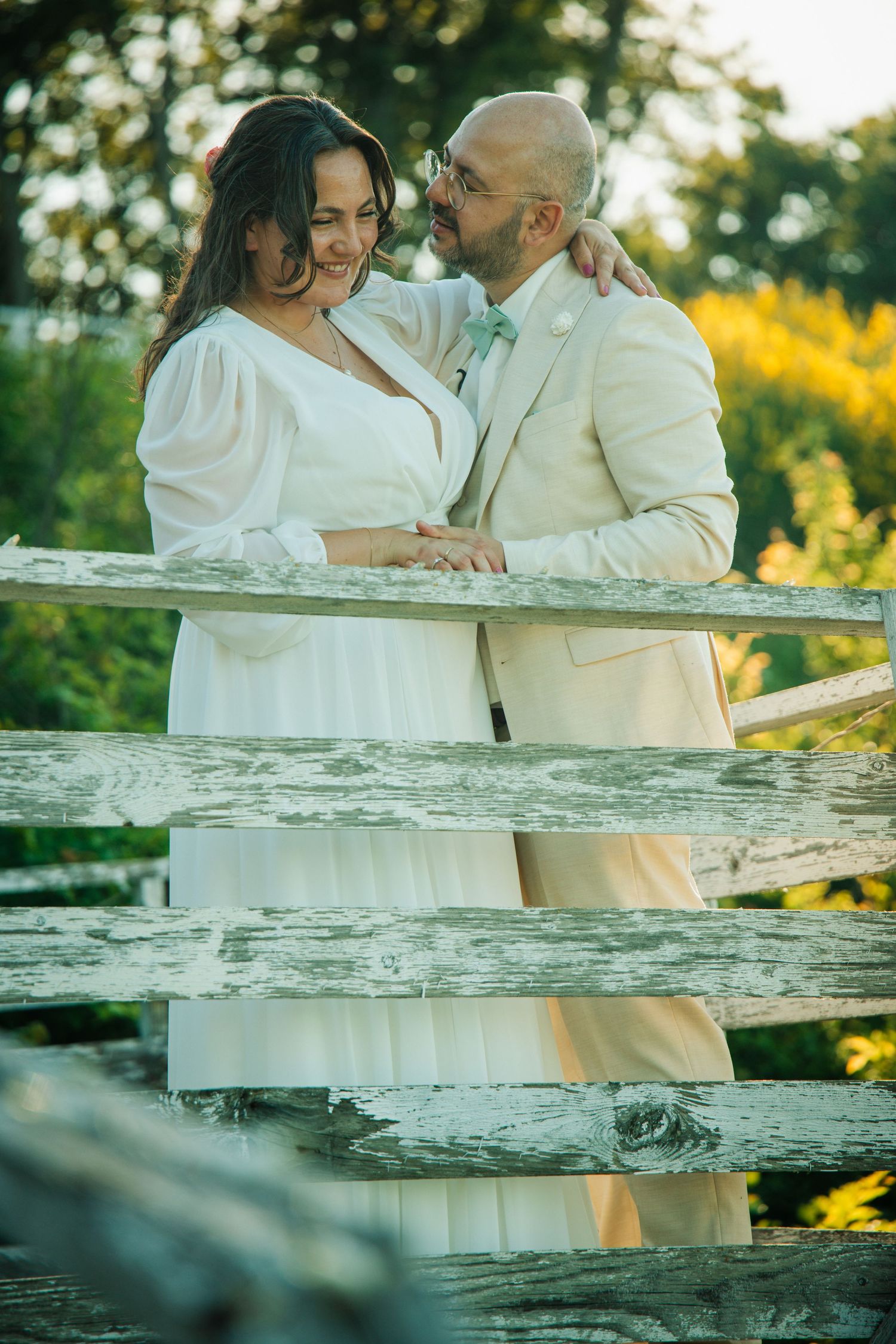Bride and groom in white attire share a romantic moment by a rustic wooden fence in a sunlit garden.