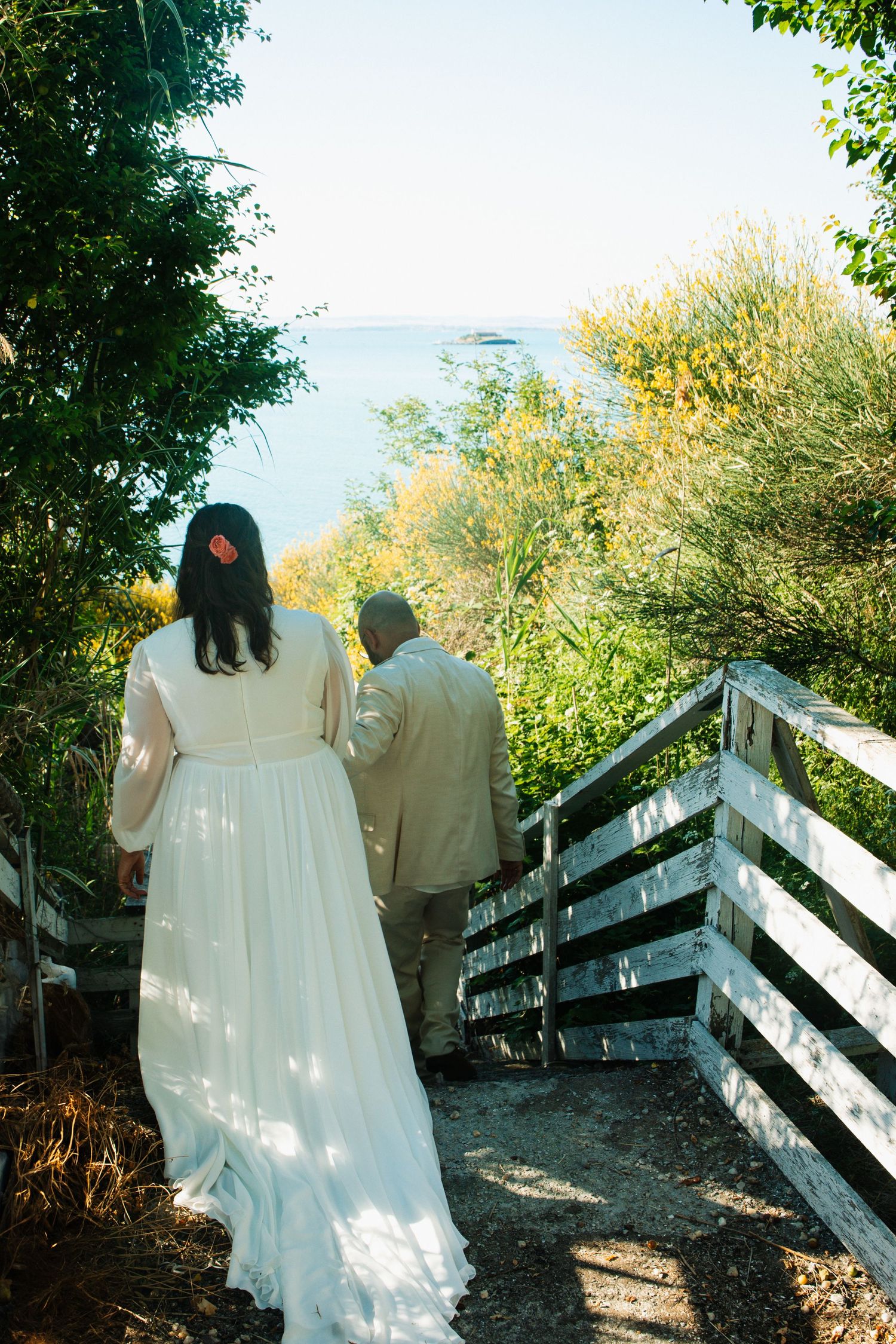 Bride and groom walking down wooden steps toward ocean surrounded by lush greenery on a sunny wedding day.