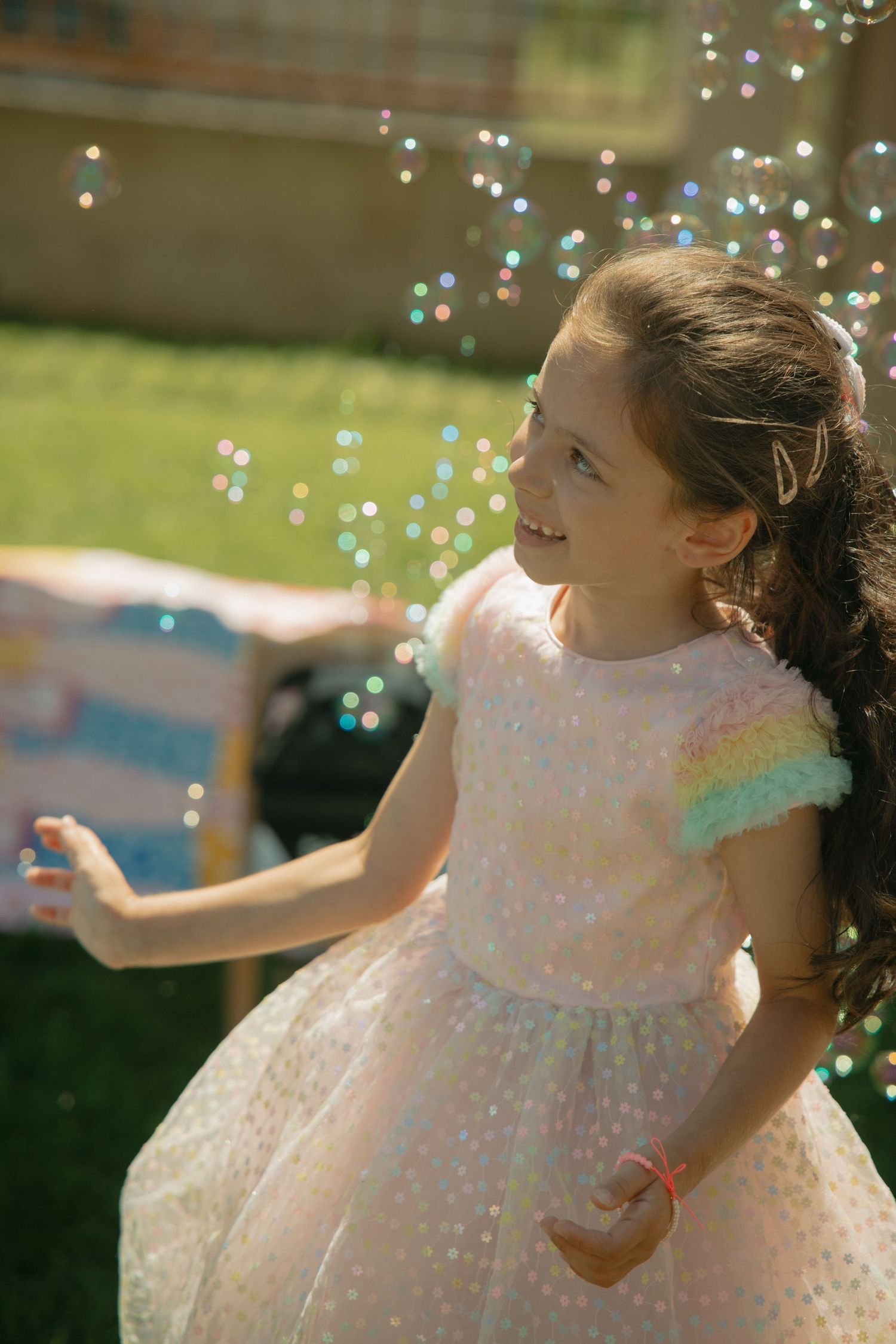 Young girl in sparkly rainbow dress playing with bubbles outdoors at a birthday party on a sunny day.