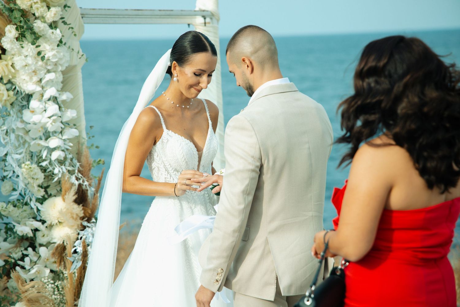 Bride and groom exchange rings at an outdoor beach wedding ceremony with white floral arch and ocean backdrop.