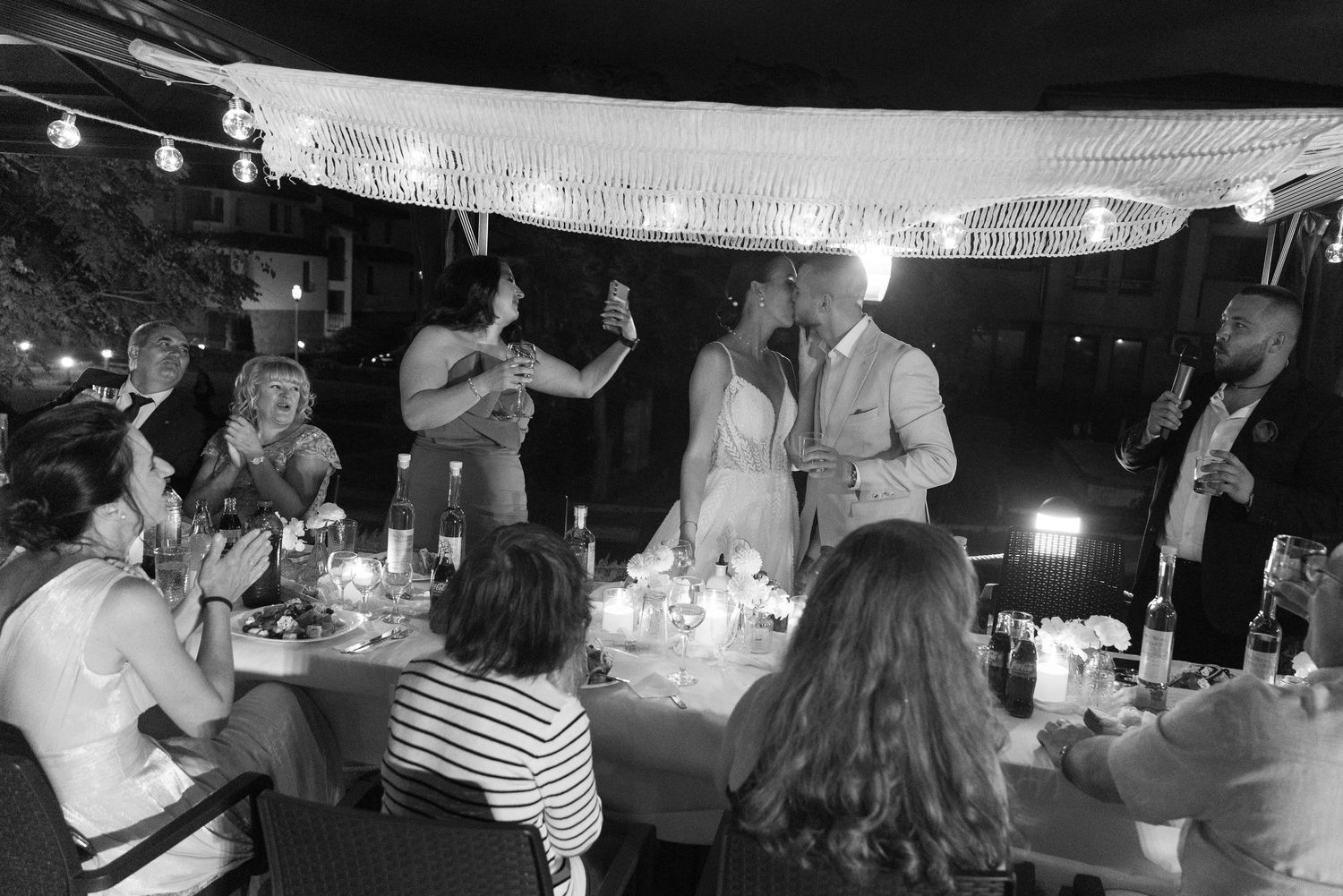 Black and white photo of wedding guests toasting the bride and groom at an elegant outdoor reception dinner.