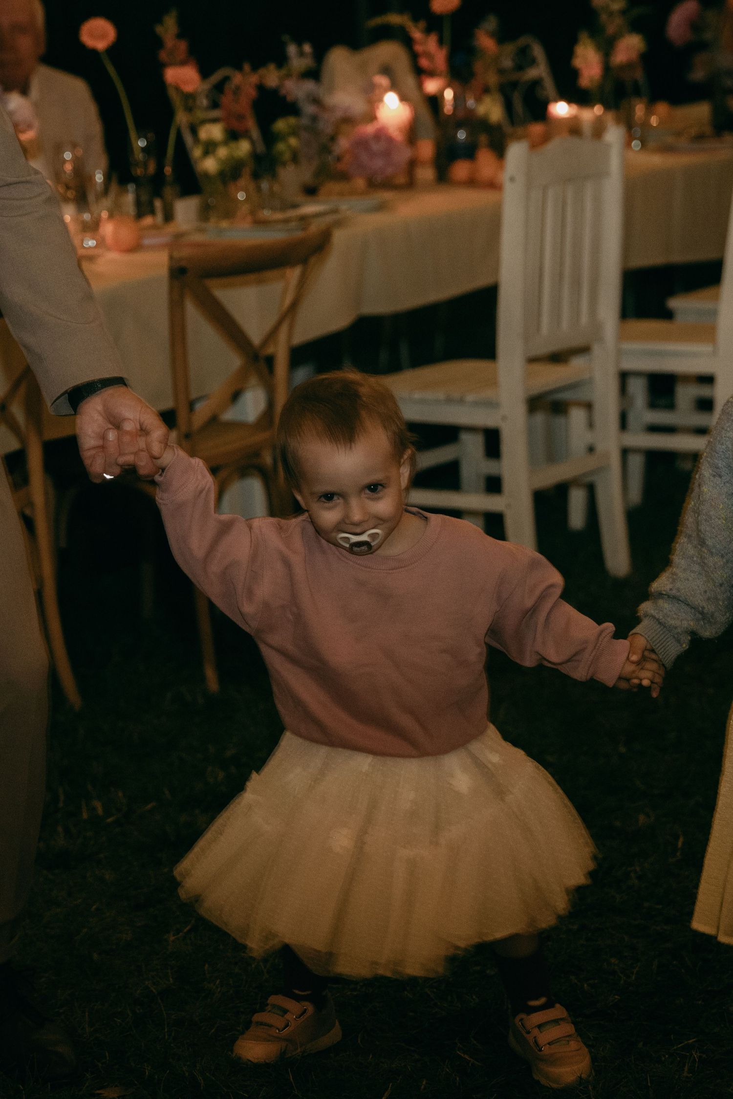 Toddler in pink sweatshirt and yellow tutu skirt holds hands with adults at an outdoor wedding reception.