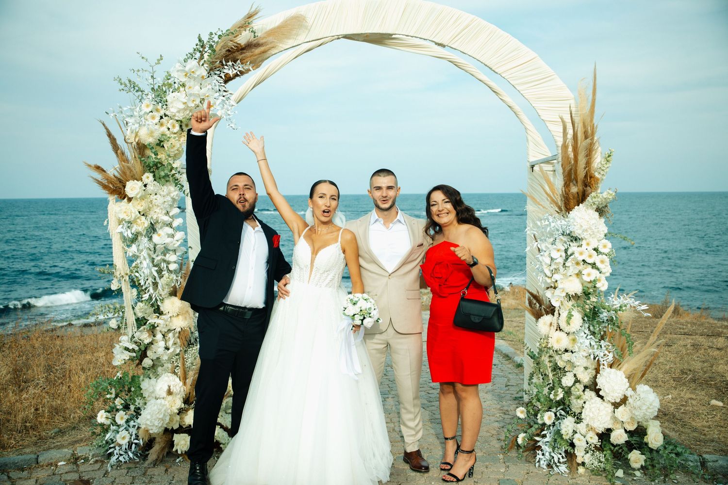 Happy wedding party of four posing under floral arch by the sea, bride in white gown and guest in red dress.