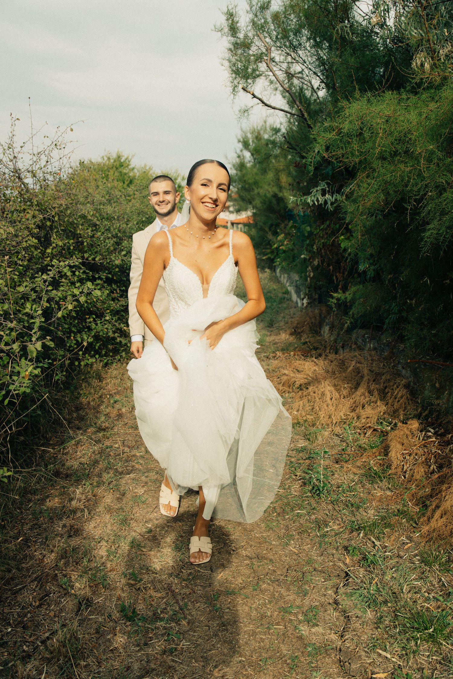 Smiling bride in white gown walks path with groom behind her, surrounded by lush greenery on their wedding day.