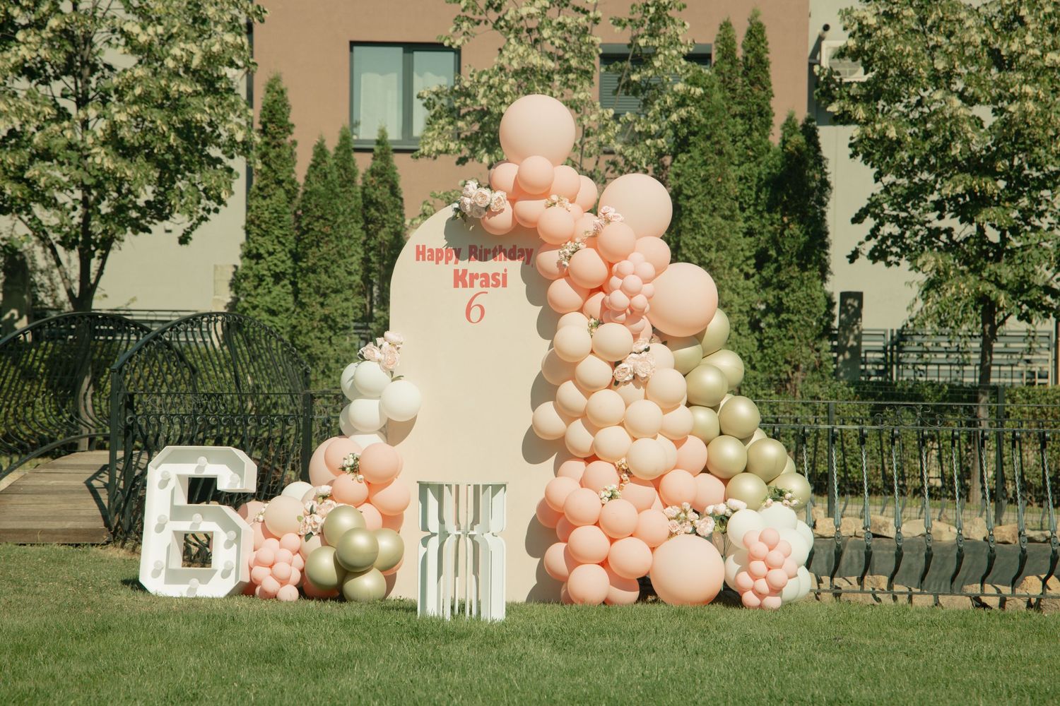 Pink and gold balloon arch decorating a 6th birthday party outdoor setup with number balloons on green lawn.