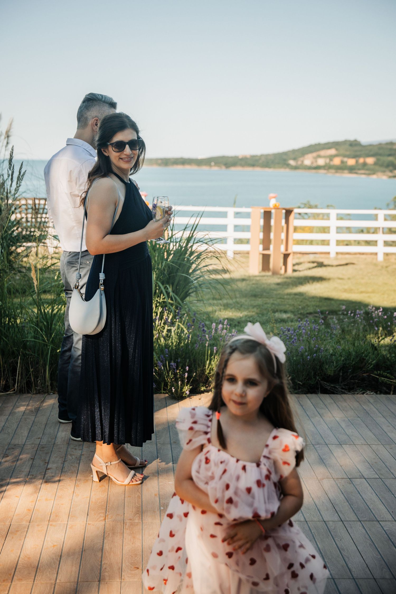 Woman in black dress with child in pink floral dress at waterfront wedding venue.