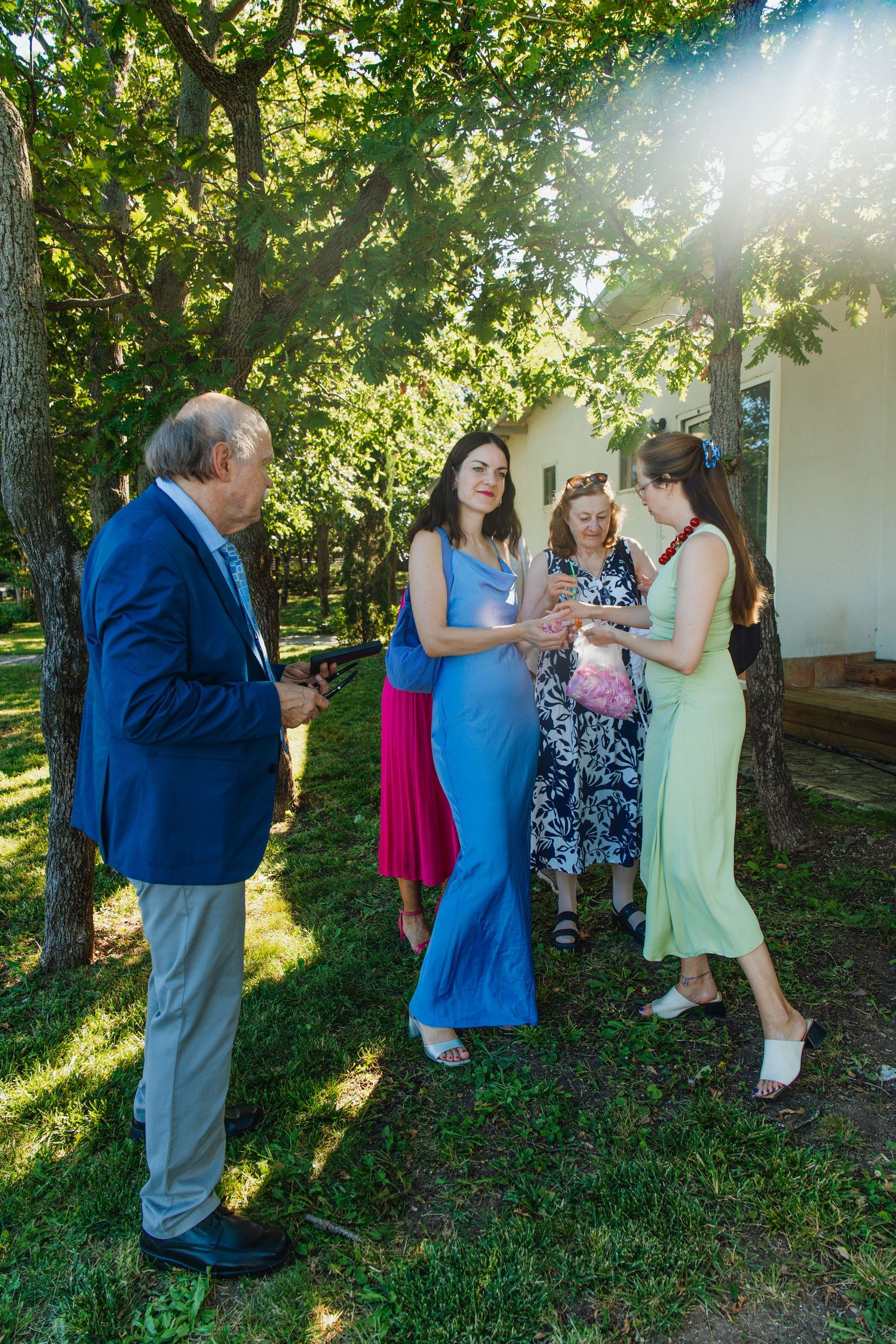 Group of elegantly dressed guests socializing outdoors under trees at a sunny wedding.