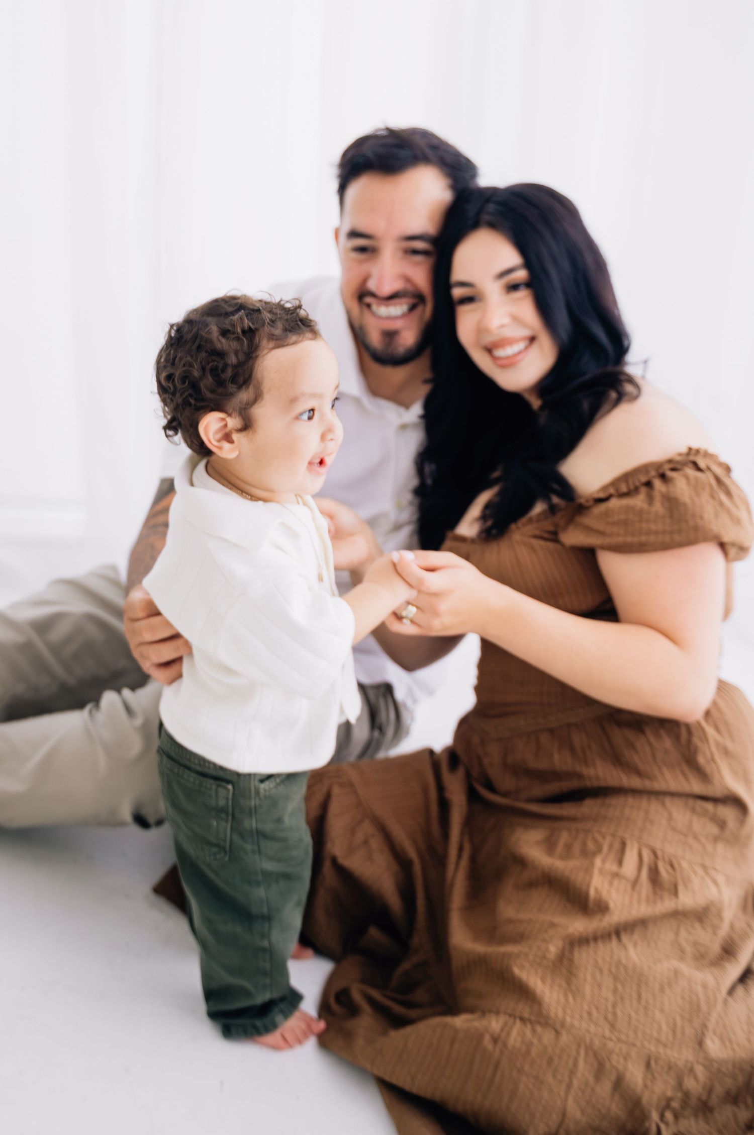 Happy family with smiling parents and curly-haired toddler playing together in a bright white photography studio.
